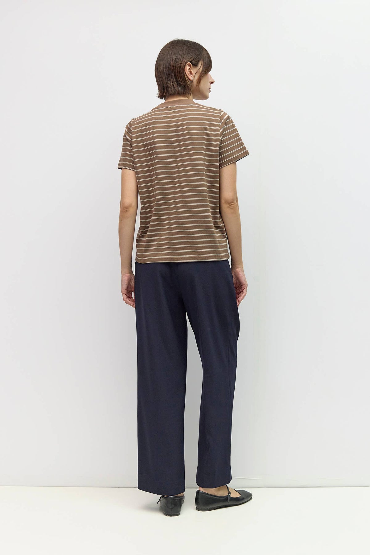 A model shown from the back wearing a brown and beige striped short-sleeve t-shirt, navy blue wide-leg trousers, and black flats, set against a plain white studio background.