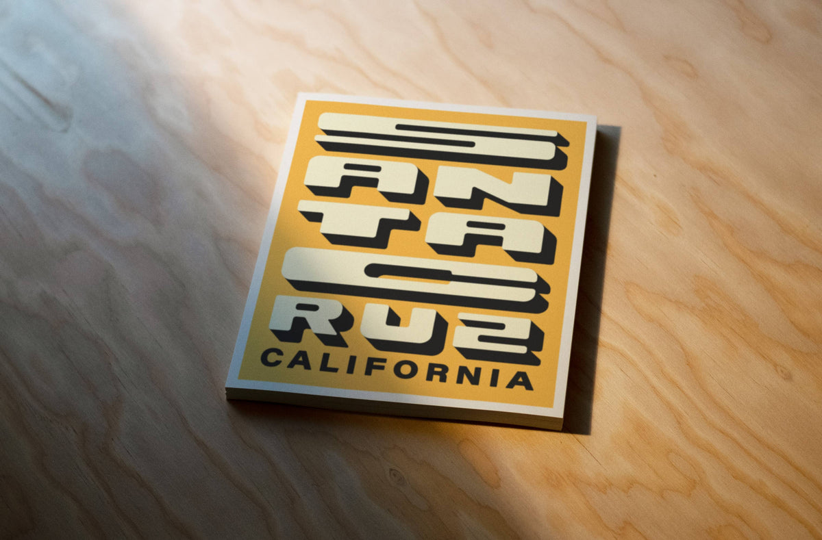 A square poster featuring the text 'Santa Cruz California' in bold black and cream lettering on a mustard yellow background, placed on a light wood surface.