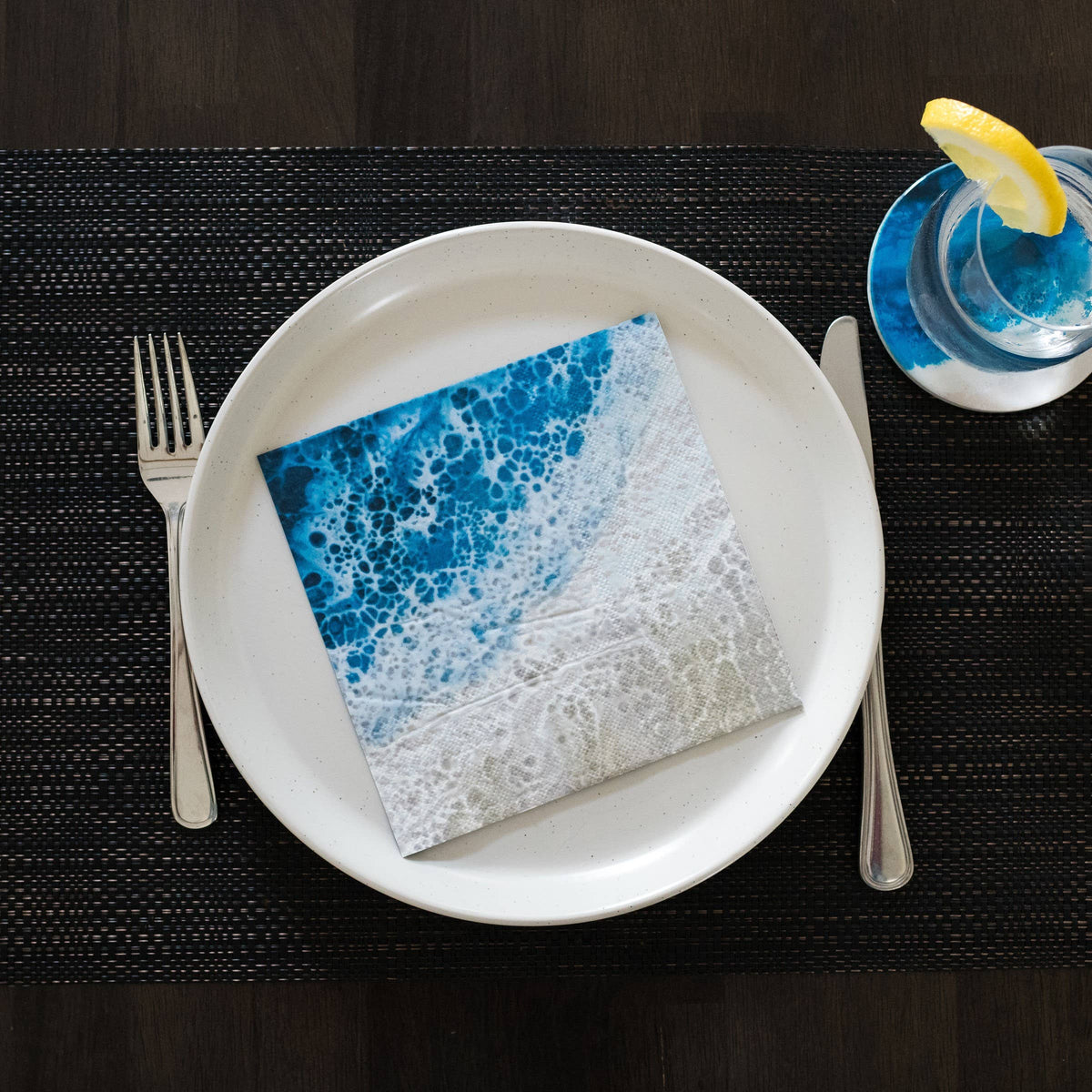 Napkin with blue and white pattern on a white plate with cutlery and a glass of water.
