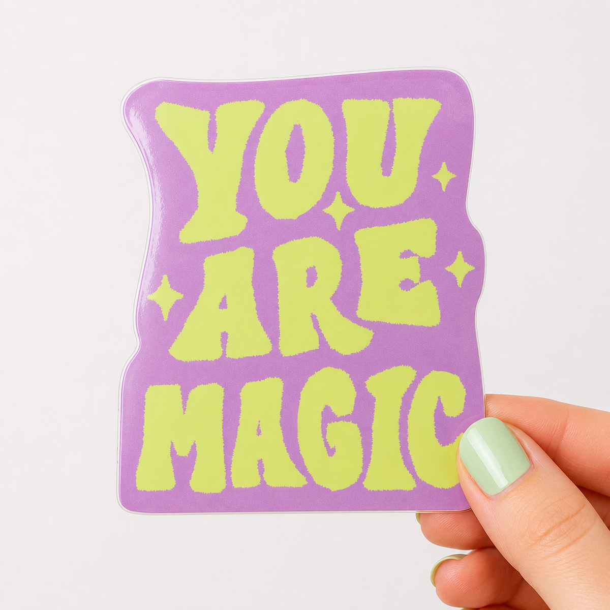 A hand with mint green nail polish holds a die-cut sticker shaped like a slice of toast. The sticker has a lavender purple background with retro-style lime green text that reads 'YOU ARE MAGIC' surrounded by small star accents.