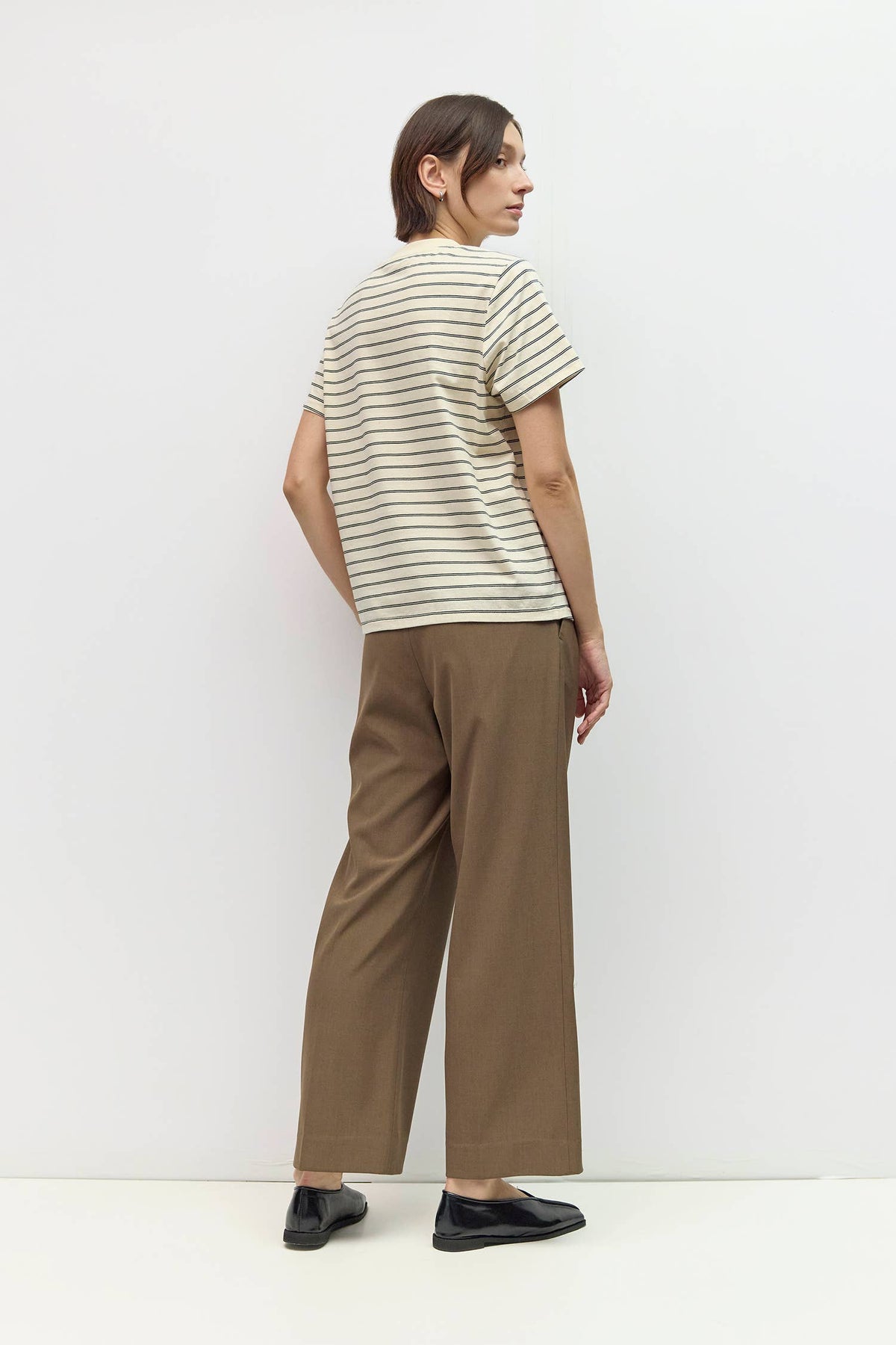 A woman is shown from the back wearing a cream-colored short-sleeve t-shirt with thin dark horizontal stripes and brown wide-leg cropped pants, paired with black shoes, against a plain white studio background.
