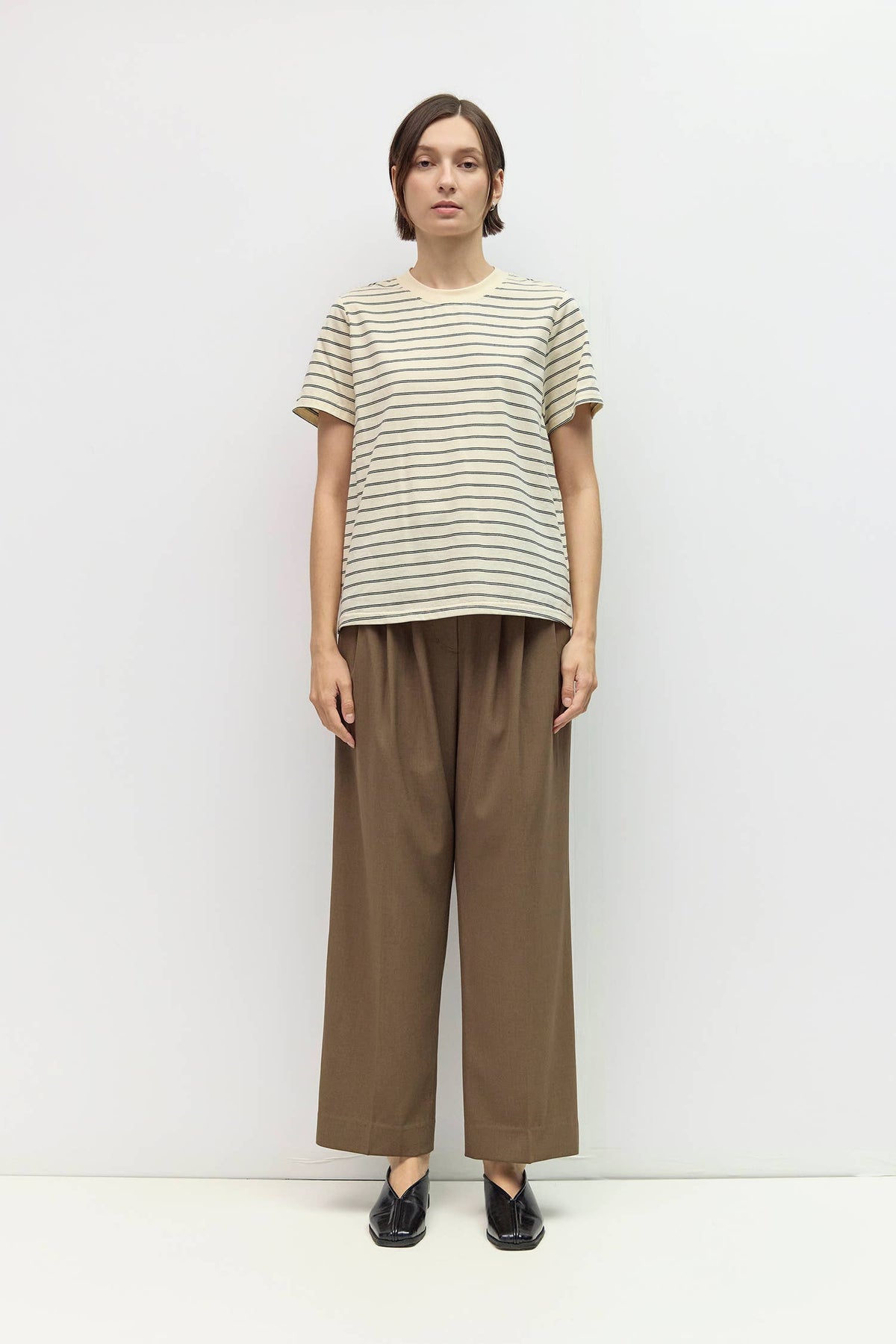 A model stands against a plain white studio background wearing a short-sleeve crewneck t-shirt with thin dark horizontal stripes on a light beige base, paired with loose brown wide-leg trousers and black shoes.