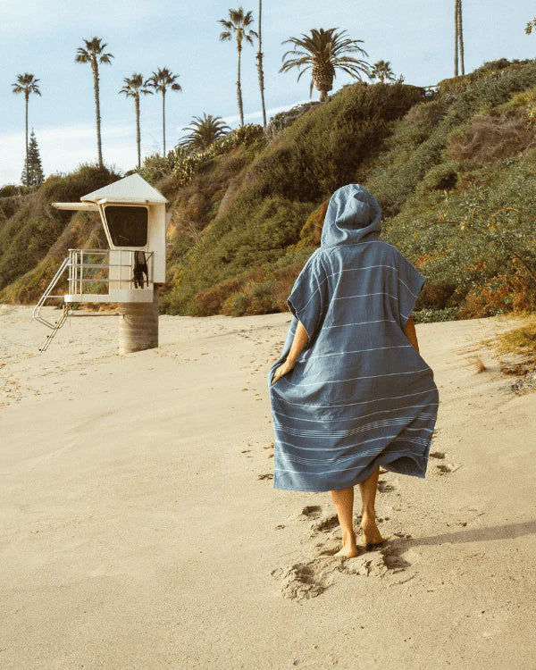 Person walking on a sandy beach with palm trees and a lifeguard tower in the background, wearing a Blue Sand Cloud Border Classic Stripe Poncho