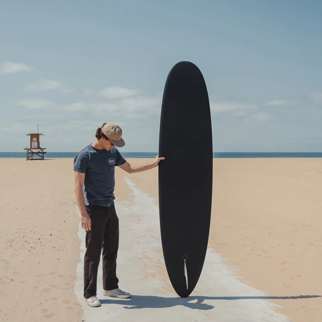Person holding a black surfboard on a sandy beach with a lifeguard tower in the background.