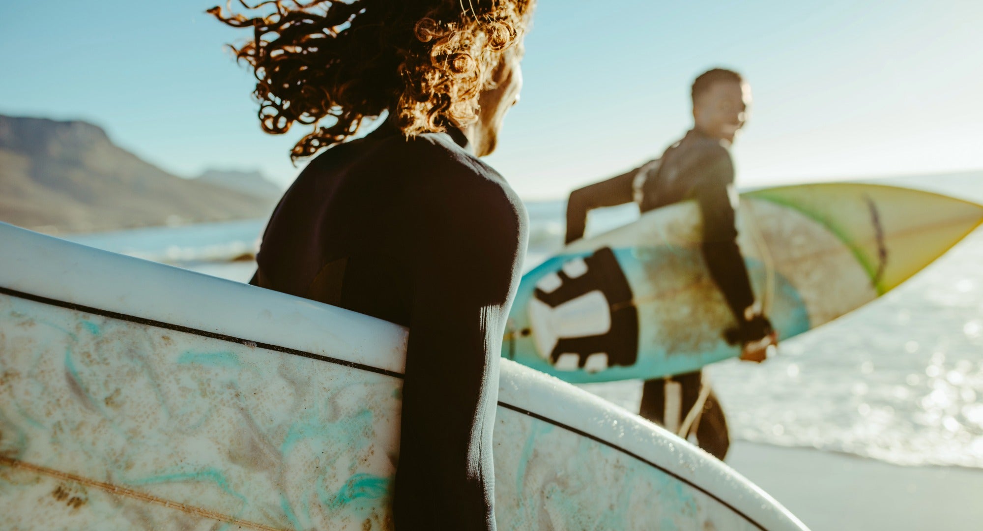 Two surfers with their boards walking on a beach with mountains in the background
