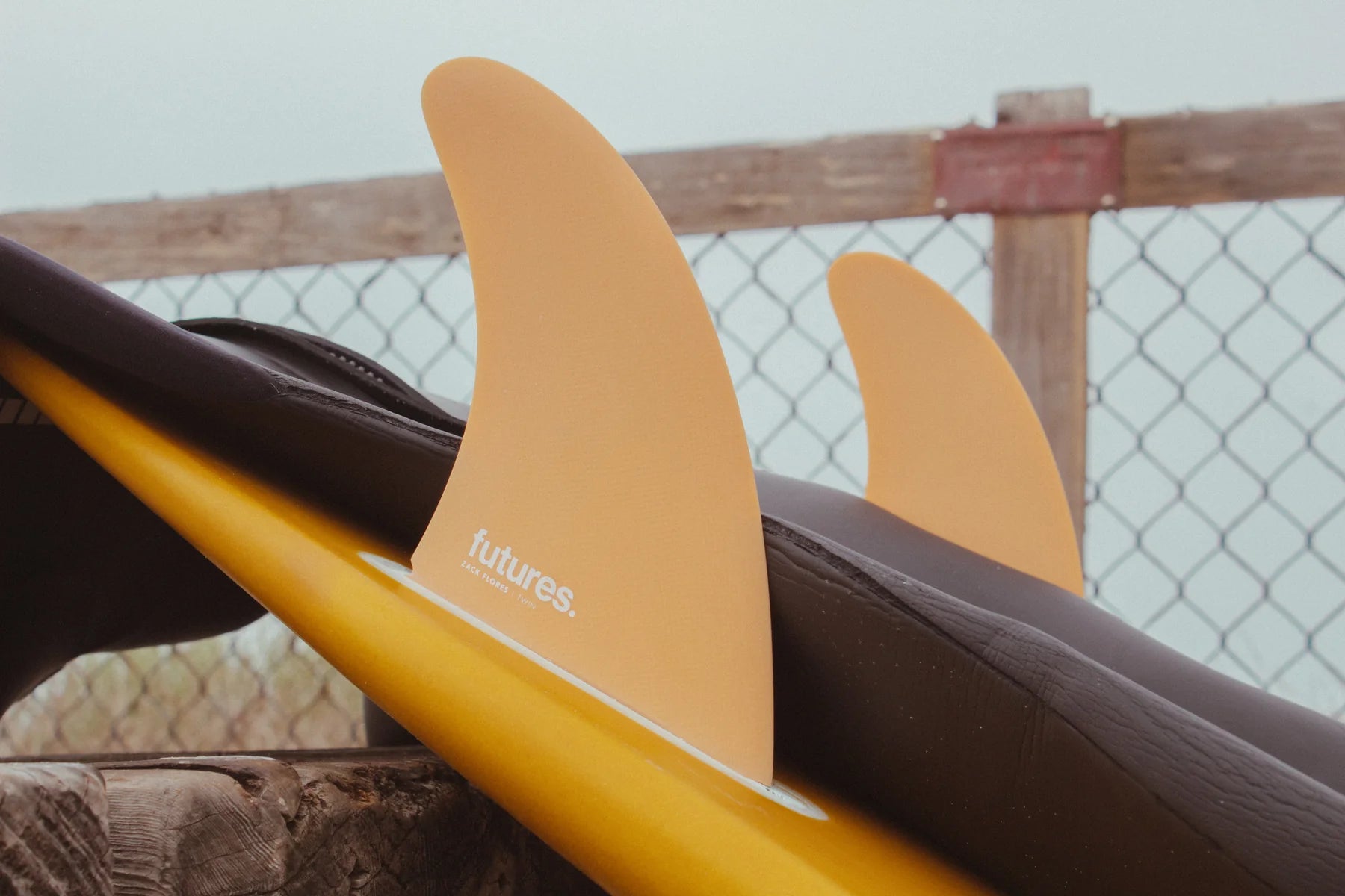 A close-up of a yellow surfboard with Futures Zack Flores Fiberglass Twin Fin Set, resting against a wooden post with a chain-link fence in the background. The fin has the brand name 'futures.