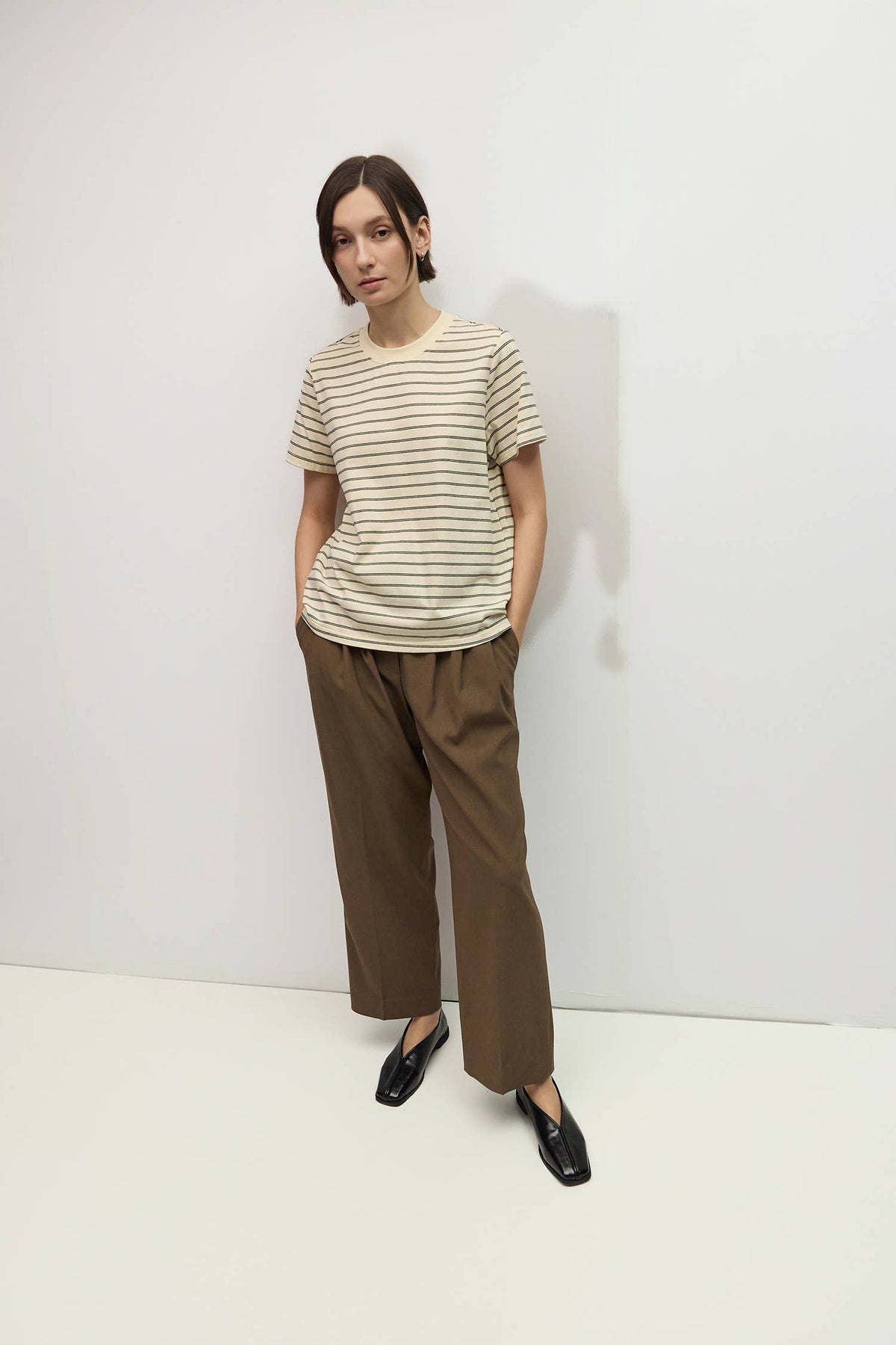A woman stands against a plain white wall wearing a cream and brown horizontally striped short-sleeve t-shirt, brown wide-leg cropped pants, and black loafers.