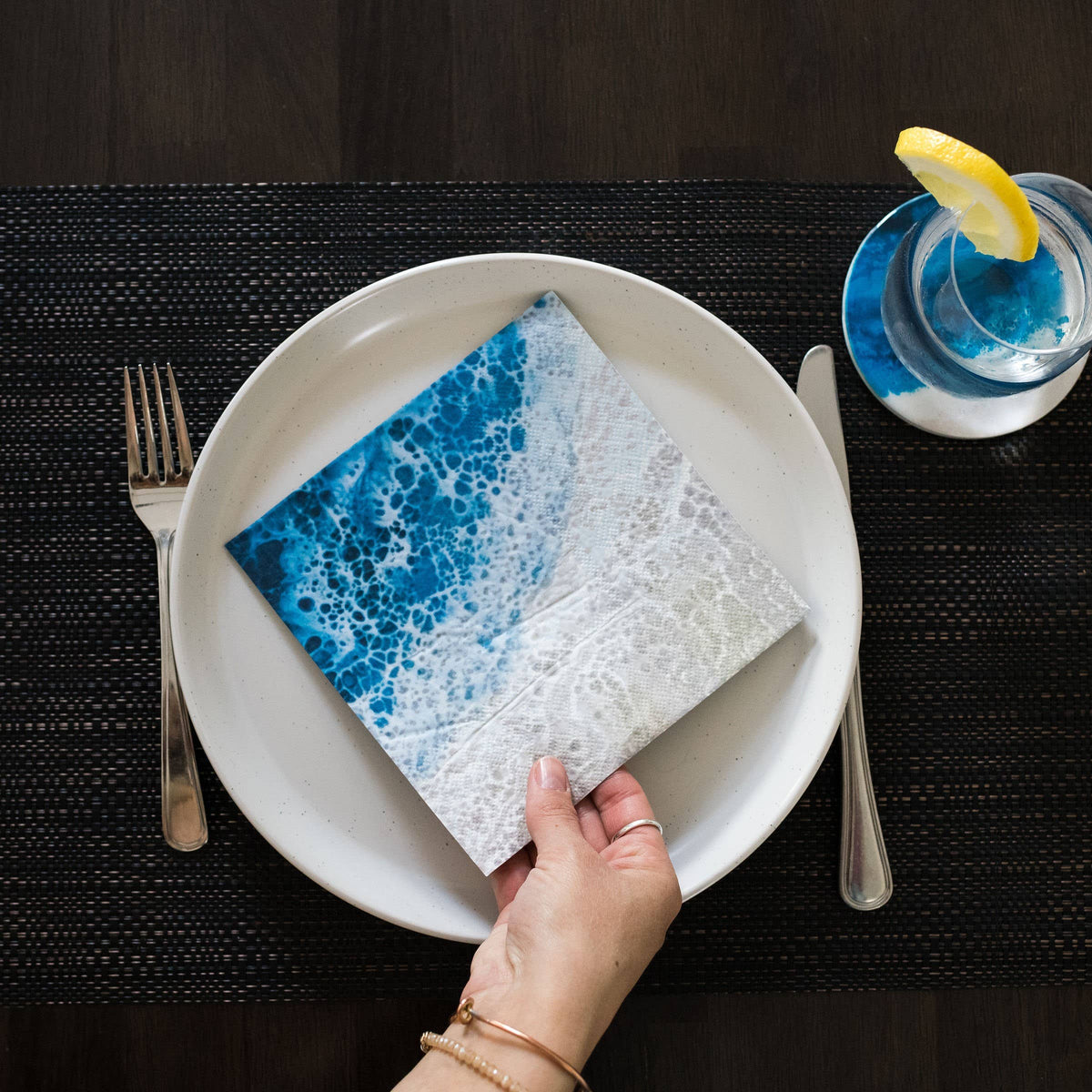 Hand placing a blue and white napkin on a white plate with cutlery and a glass of lemonade.