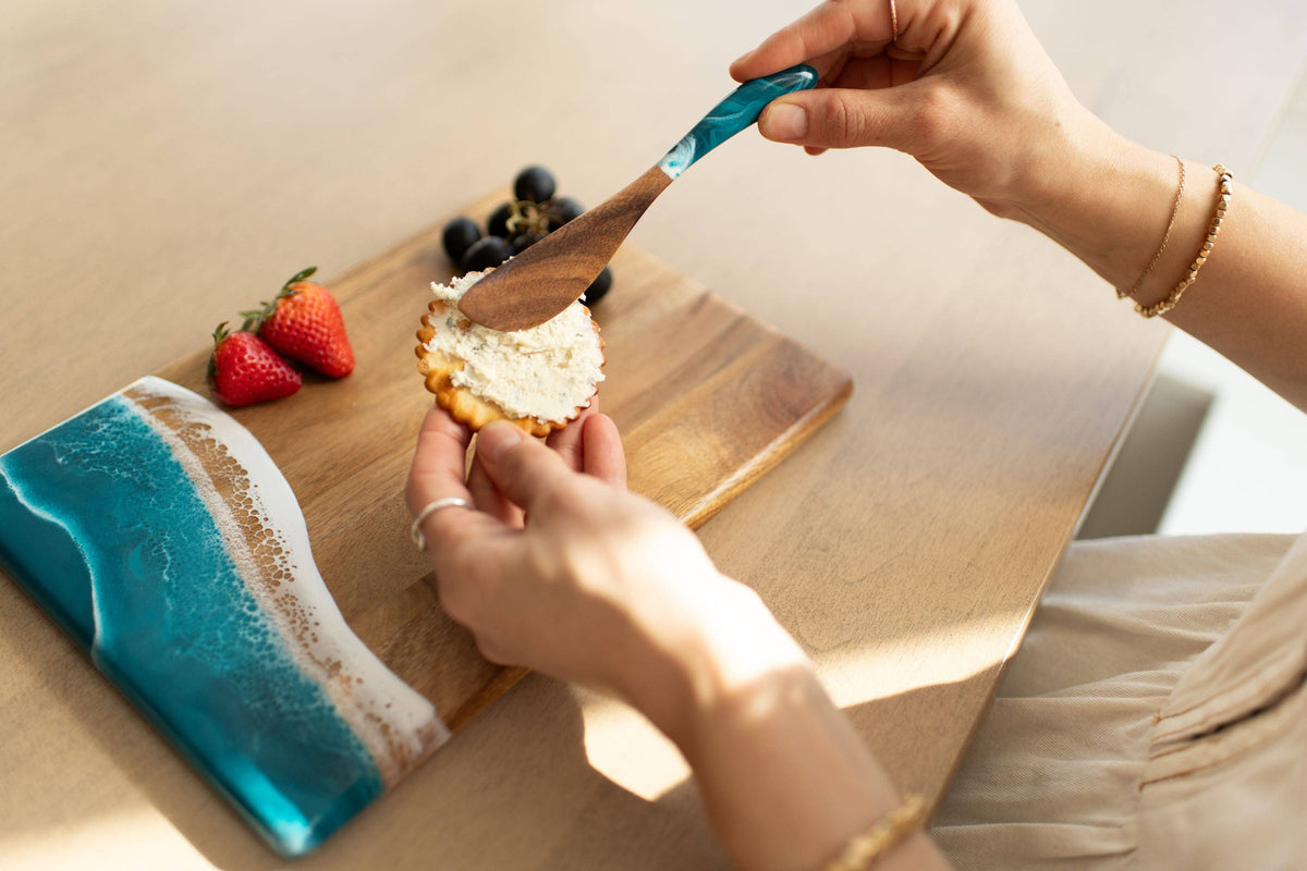 Person spreading cream on a cracker with a wooden spoon on a wooden board.