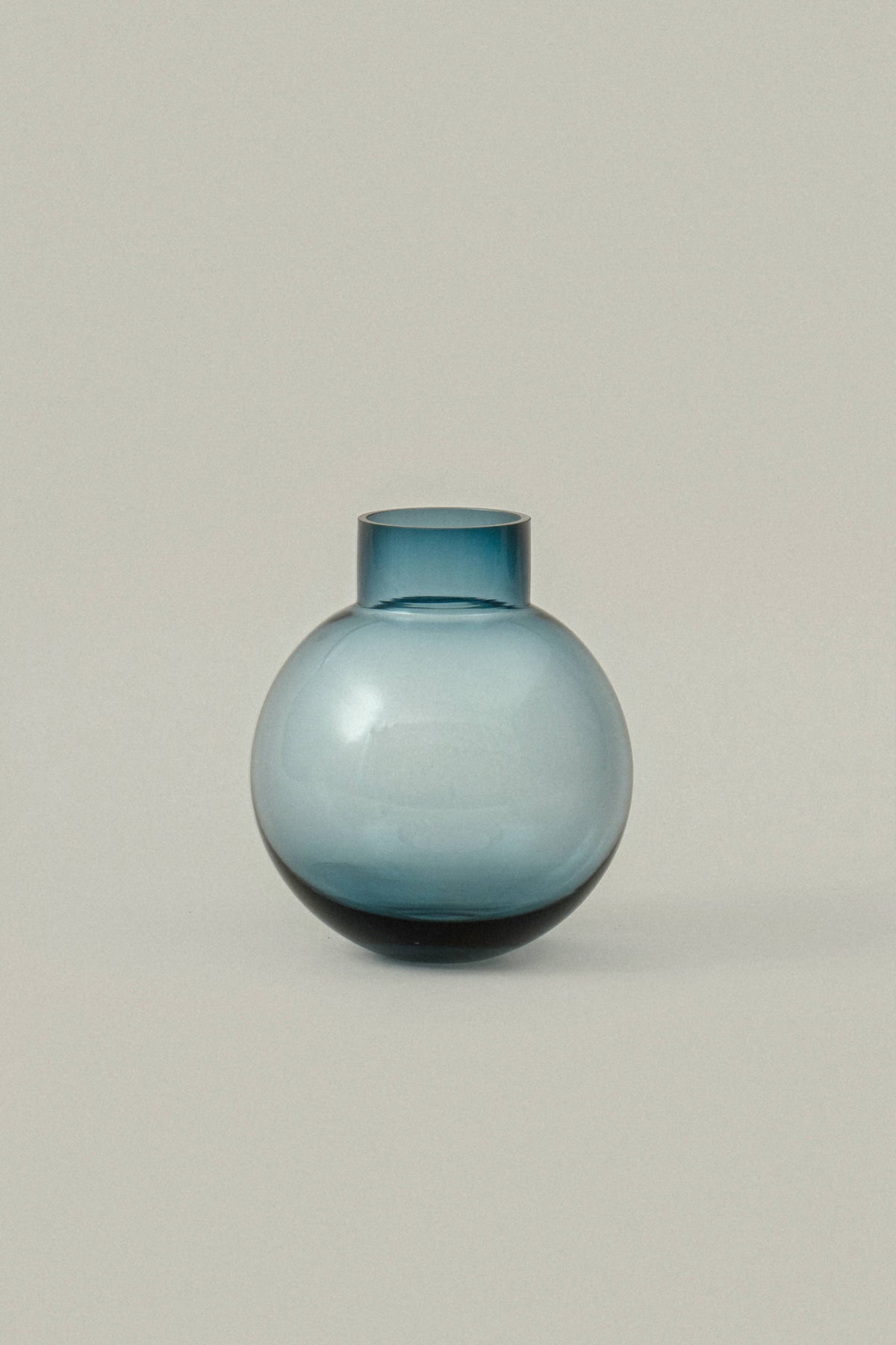 A small round glass vase with a short neck, featuring a gradient color transition from light blue at the top to dark blue at the base, set against a solid light gray background.