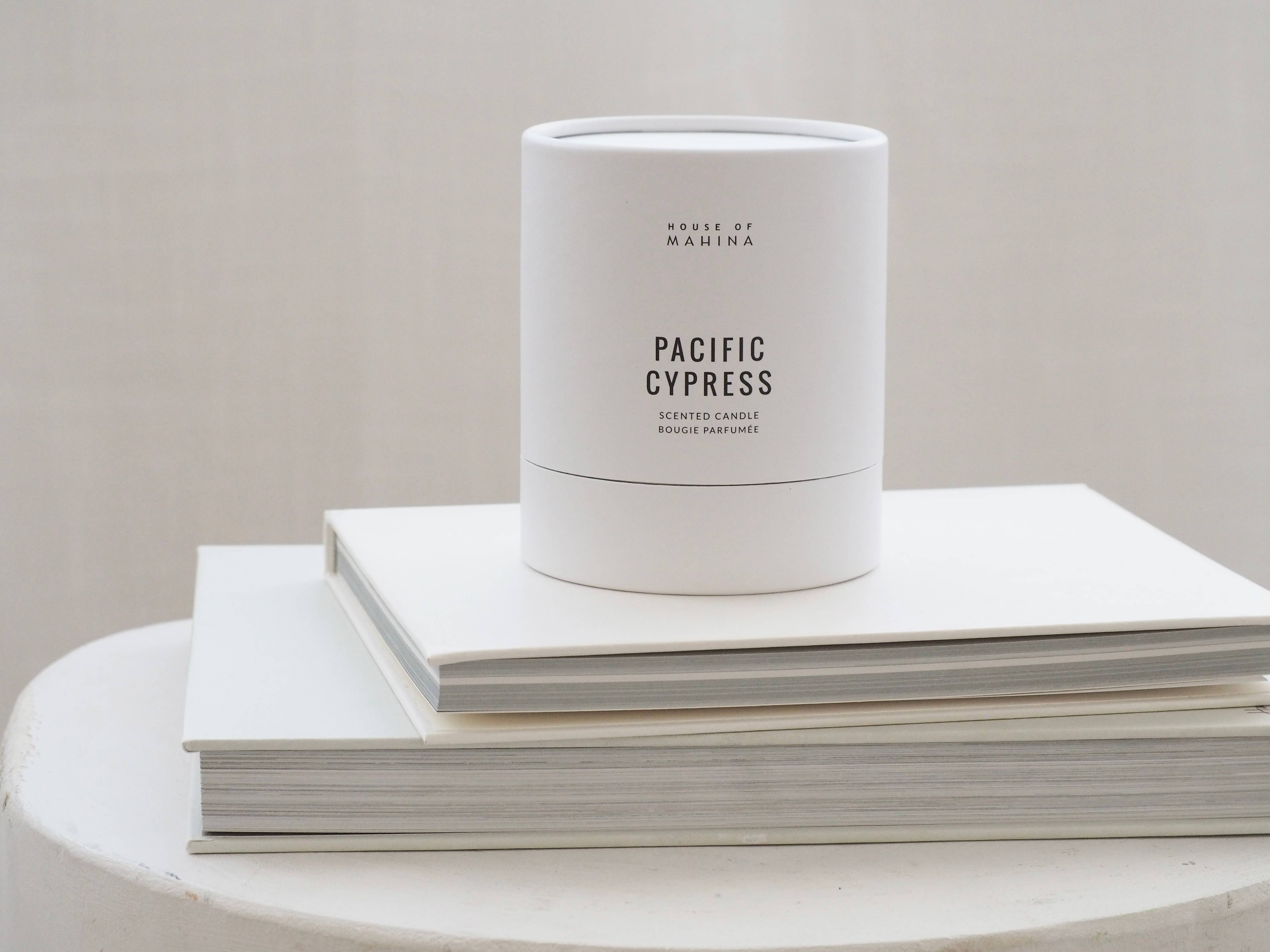 White candle labeled 'Pacific Cypress' on a stack of white books with a plain background