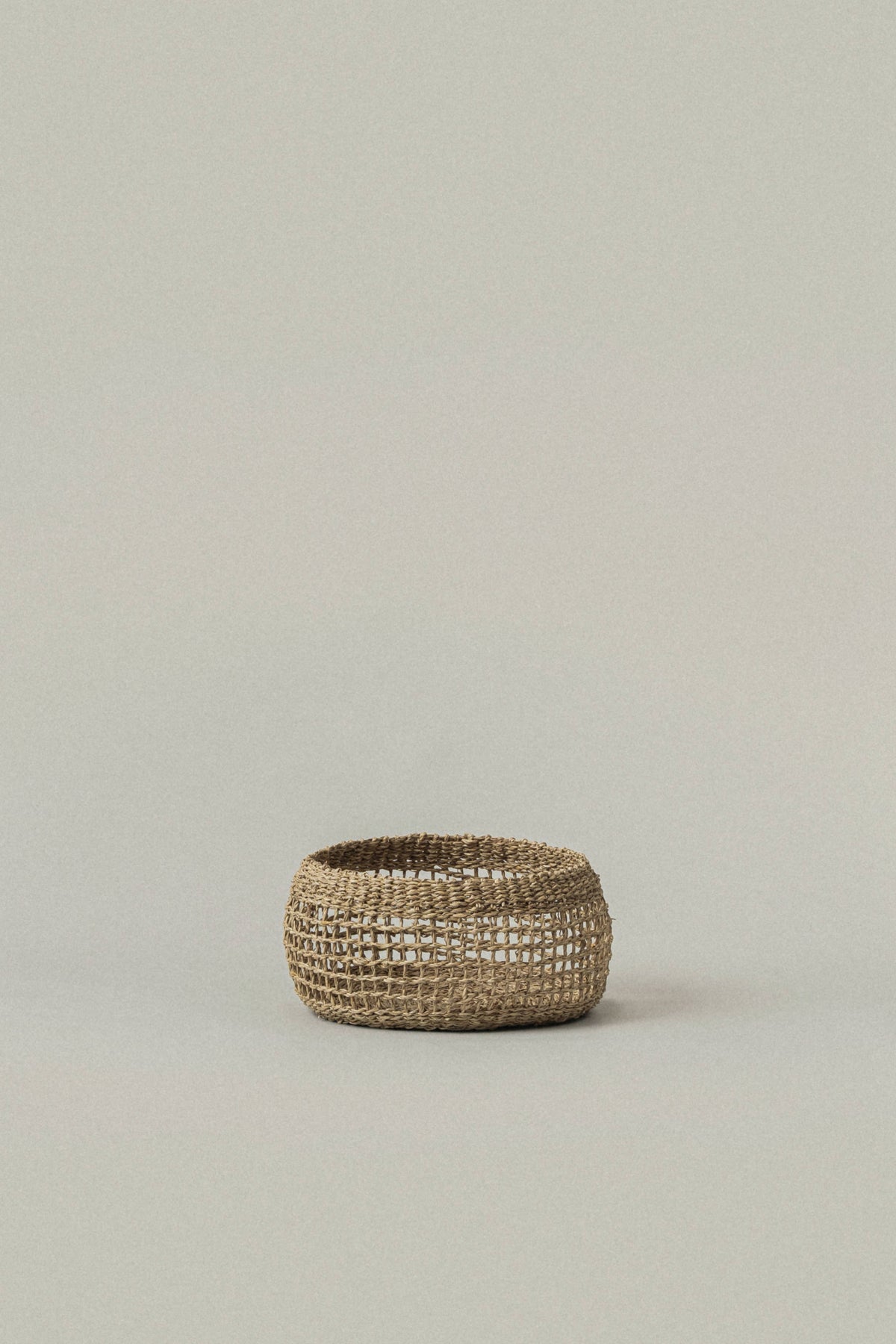 A small woven basket or planter made from natural straw fibers, shown against a solid, neutral light gray background.