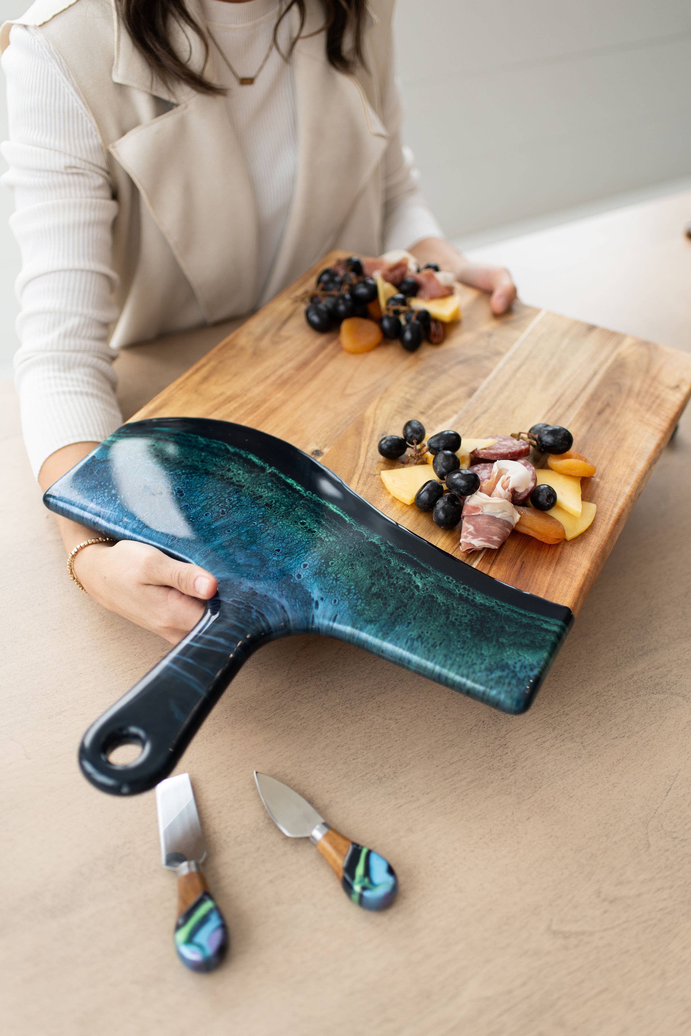 Person holding a blue cheese knife over a wooden cutting board with fruits and meats.
