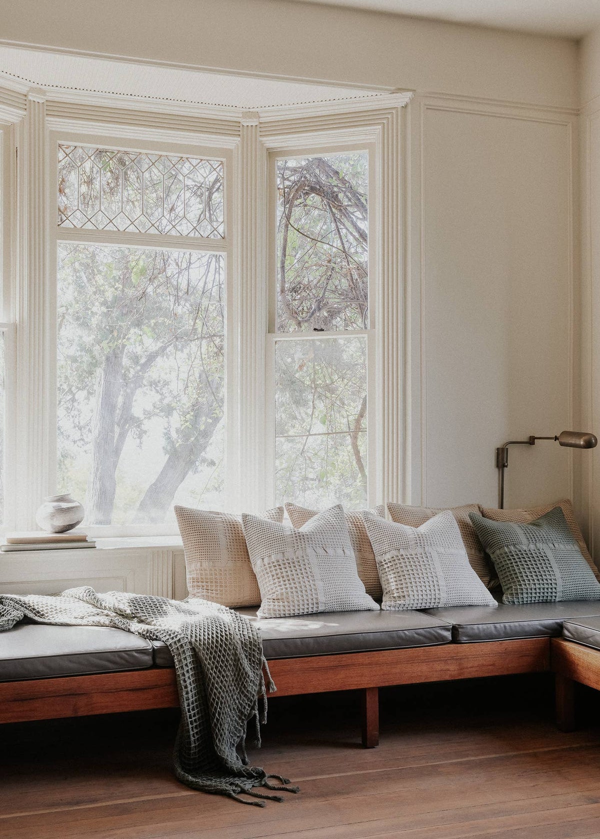 A cozy built-in wooden window seat with gray cushions, styled with multiple textured throw pillows and a gray knit blanket, set against a warm cream paneled wall with a bay window.