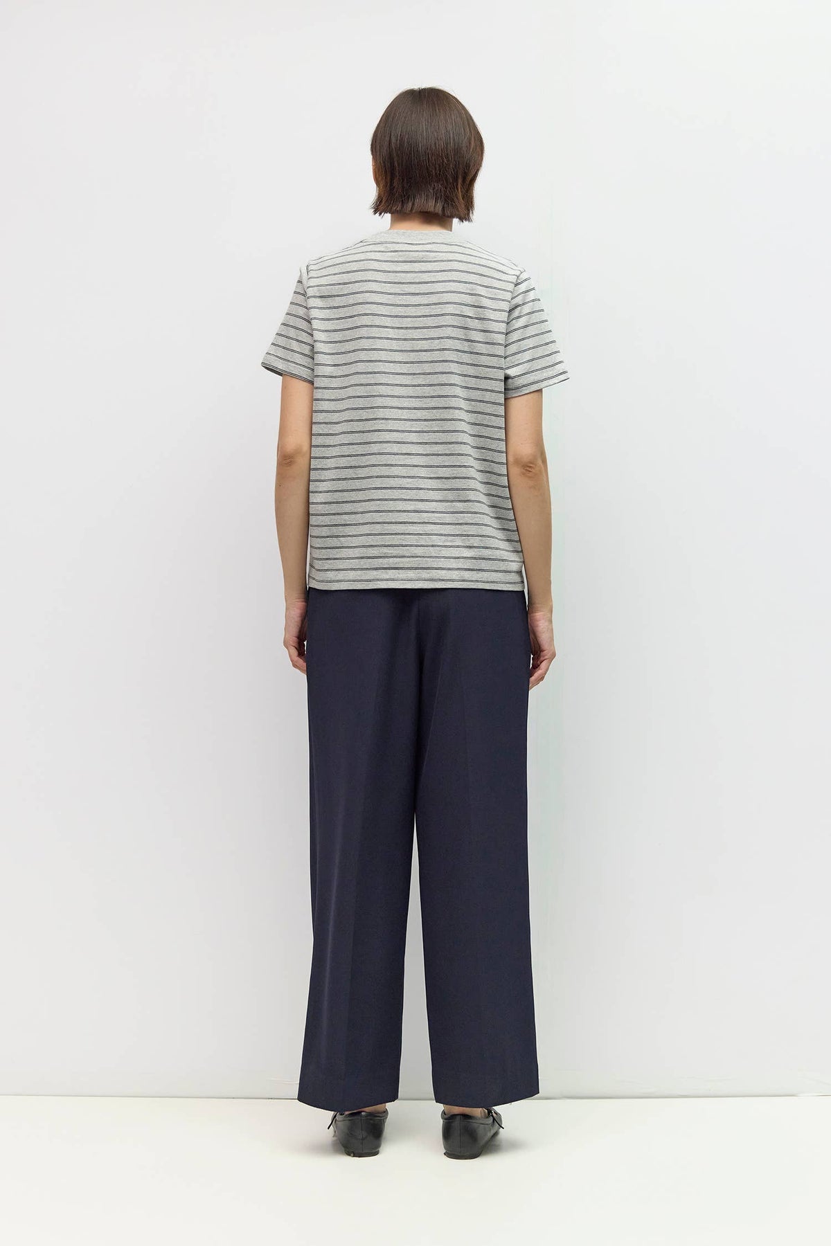 A person standing with their back to the camera, wearing a light gray short-sleeve t-shirt with thin dark horizontal stripes and dark navy wide-leg trousers, against a plain white studio background.