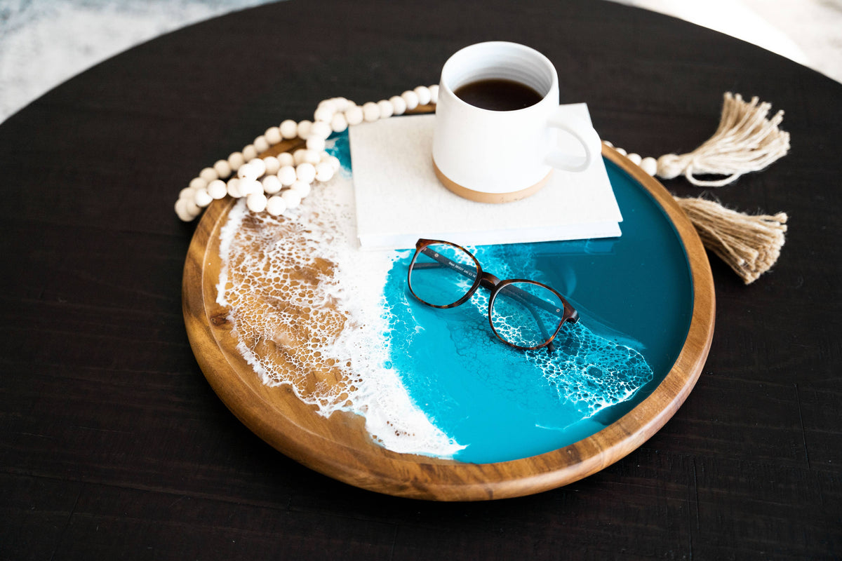 A round wooden tray with a turquoise ocean-wave design and white foam details, styled on a dark table with a white mug, eyeglasses, a beaded necklace, a napkin, and tassels.