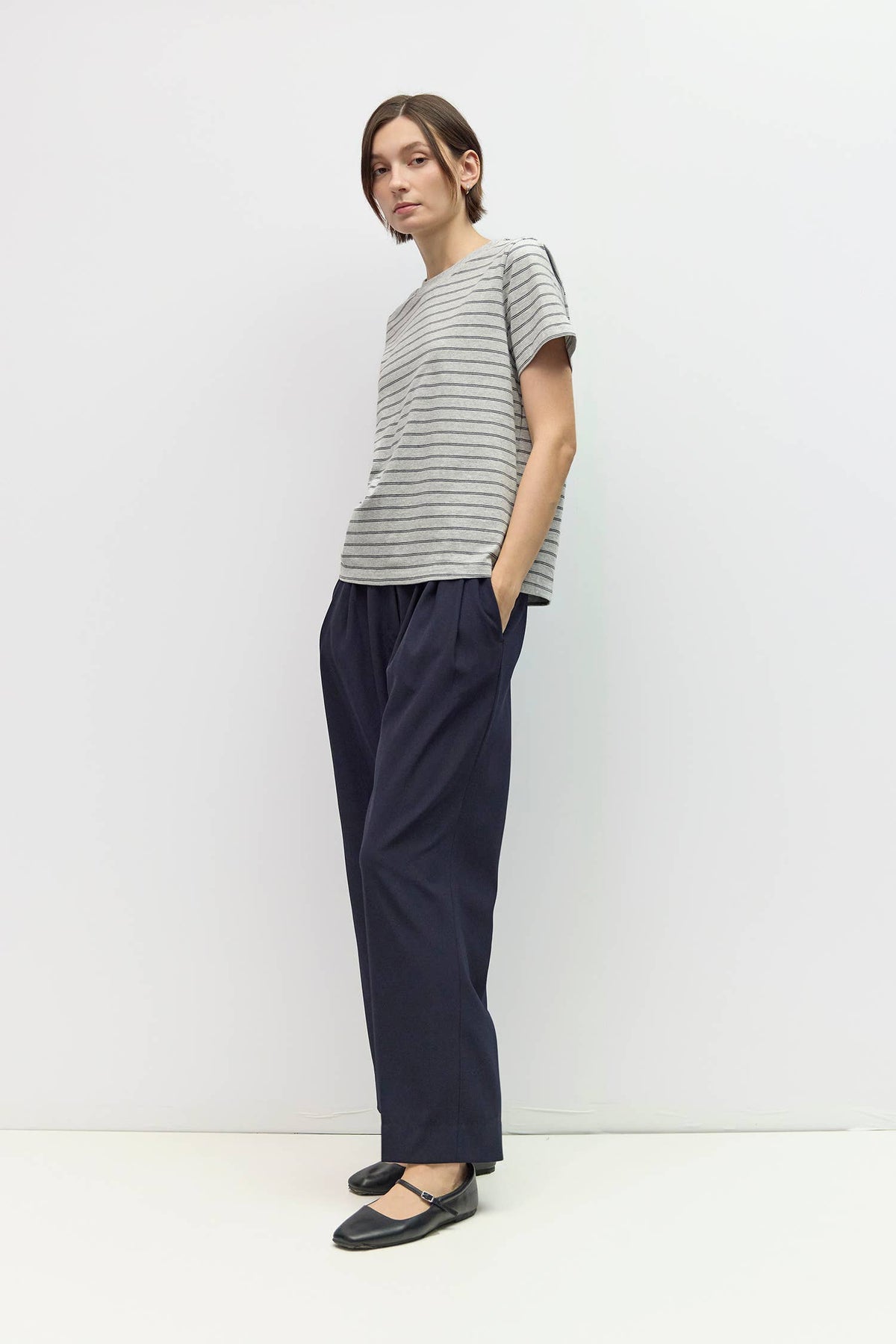 A model stands in a studio wearing a gray and white horizontally striped short-sleeve t-shirt, navy blue wide-leg trousers, and black Mary Jane flats.
