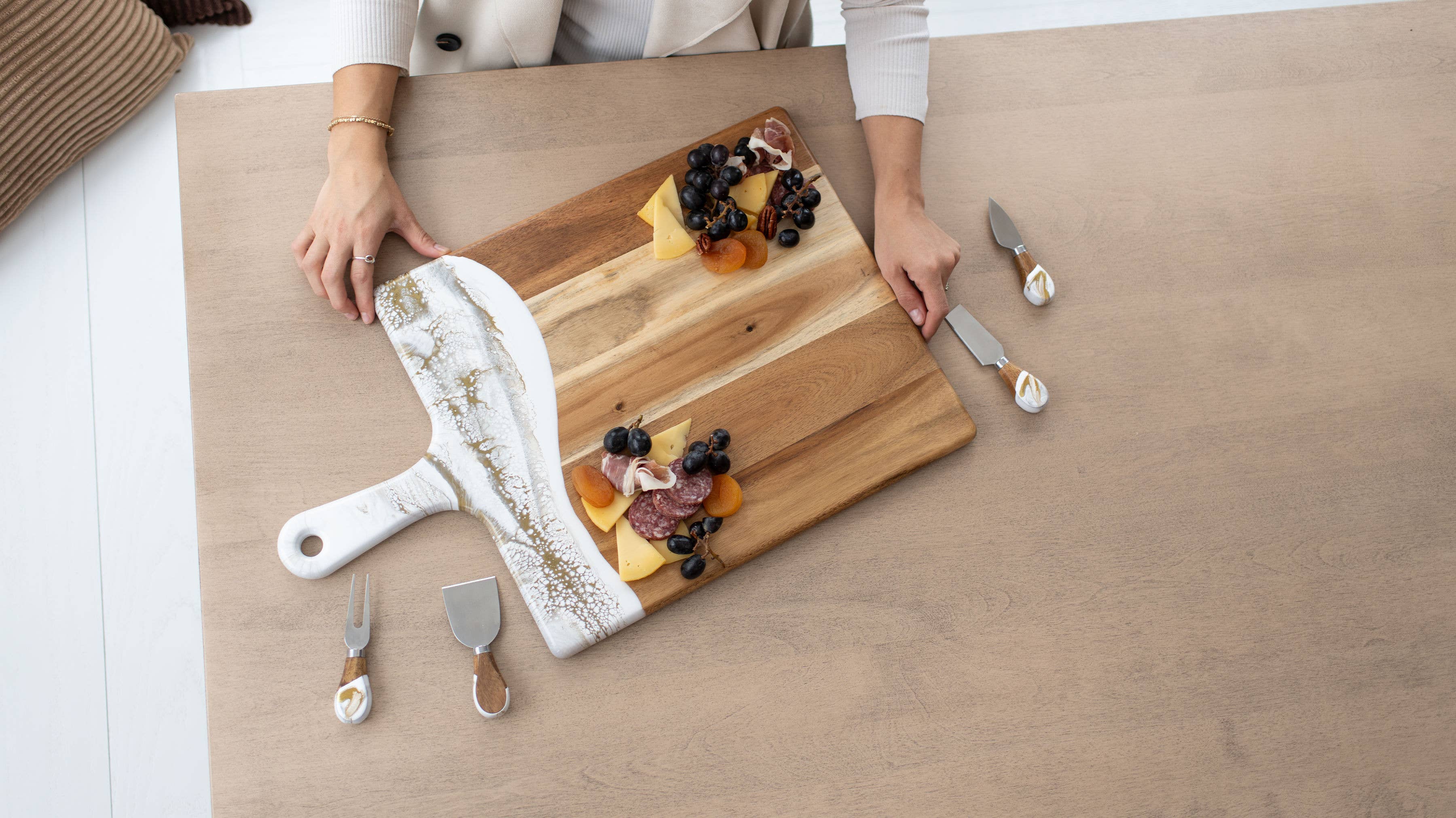Person arranging food on a wooden cutting board with utensils on a light wood surface.