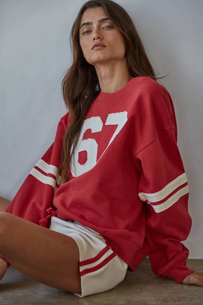 woman sitting against the wall with a red sweater on. The number 67 sewn on in contrasting colors to match the sweat shorts. 