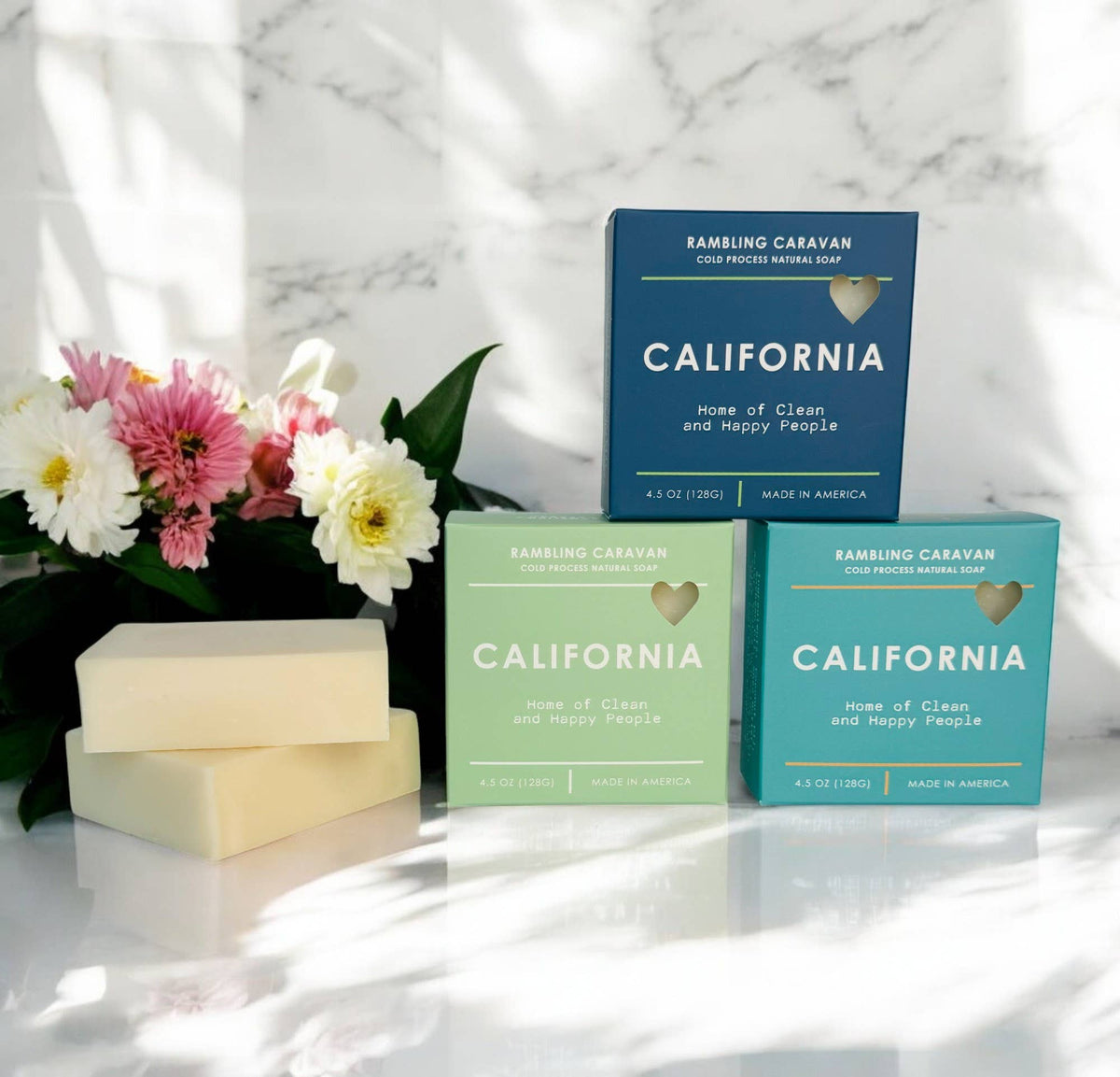 Three rectangular bars of RAMBLING CARAVAN California cold sore cleansing soap in pastel blue, mint green, and teal packaging, displayed on a white marble surface next to a bouquet of pink, white, and yellow flowers.