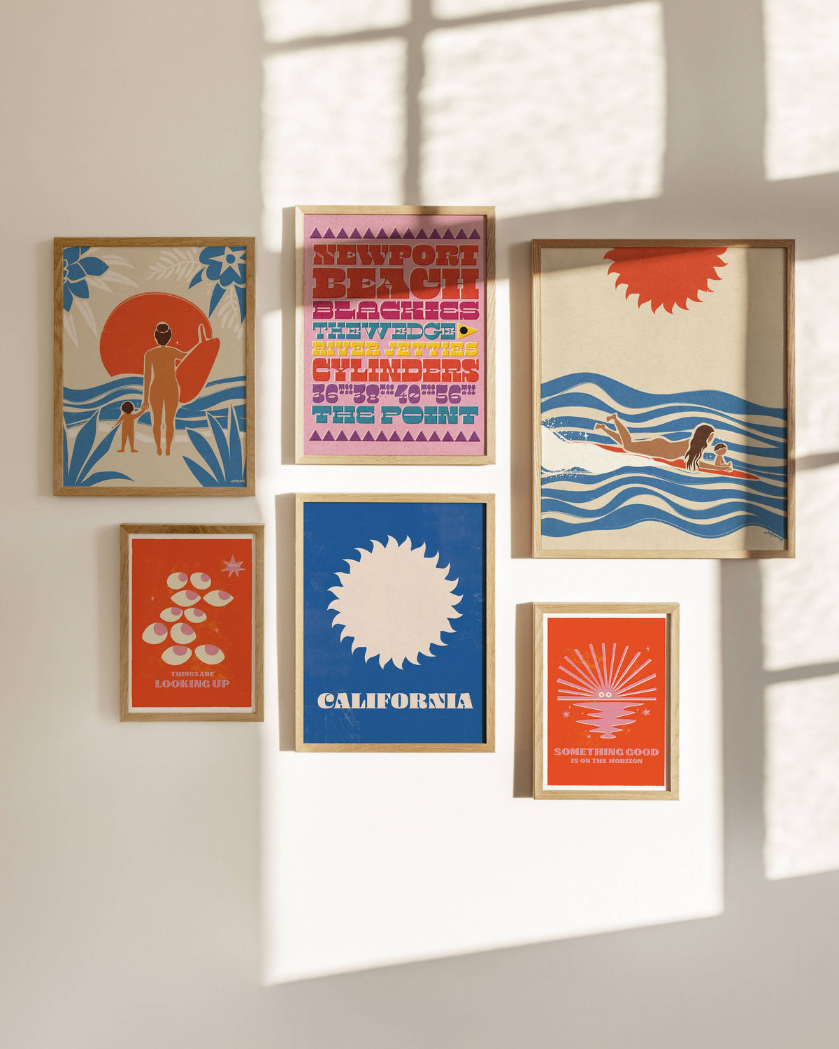 Five framed art prints with a retro California beach theme are arranged in a gallery wall under natural window light with soft shadows. The prints feature bold colors like blue, orange, pink, and cream, with illustrations of a surfer, palm trees, waves, and a sun.