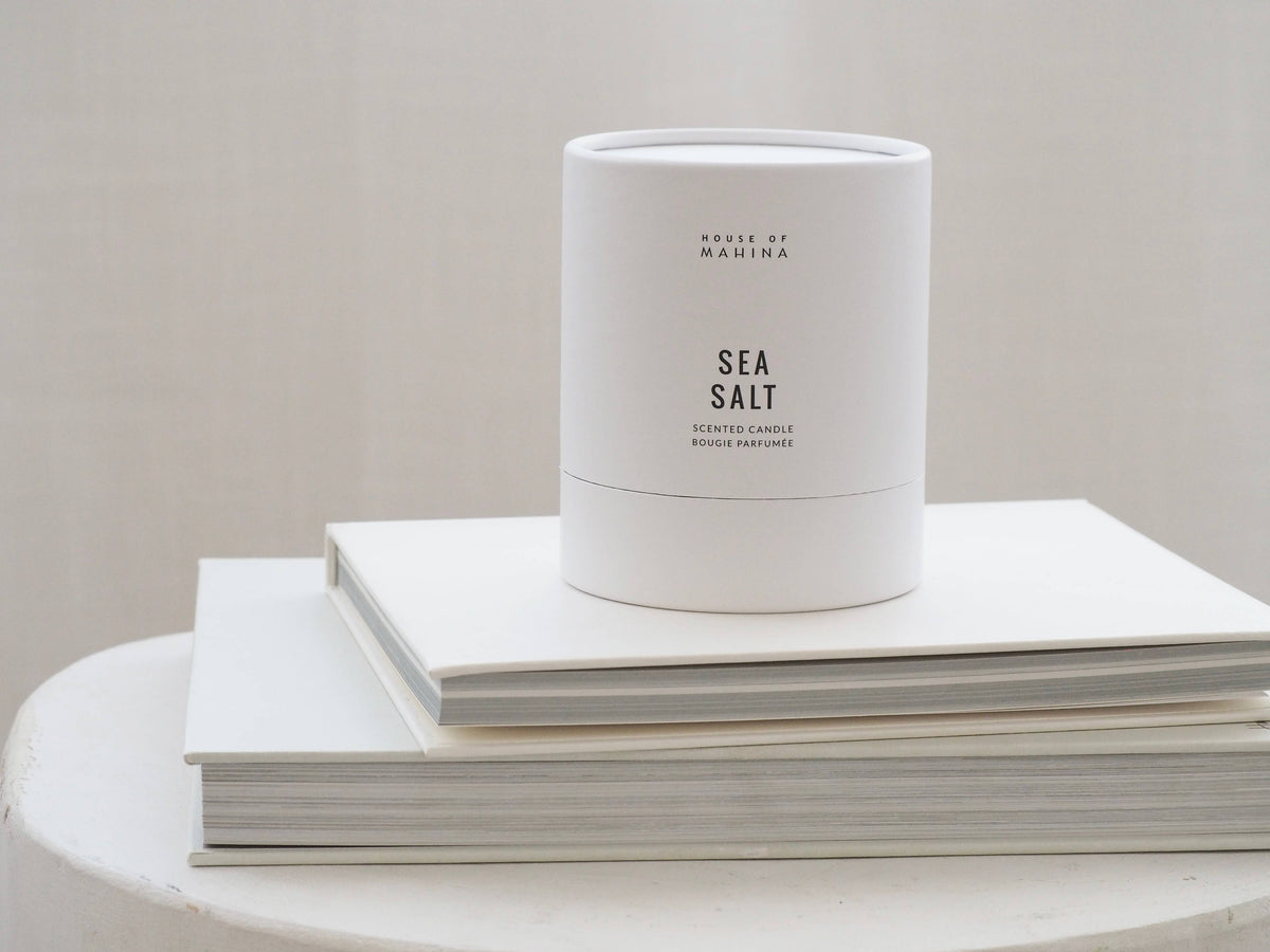 White candle labeled 'Sea Salt' on a stack of white books with a plain background