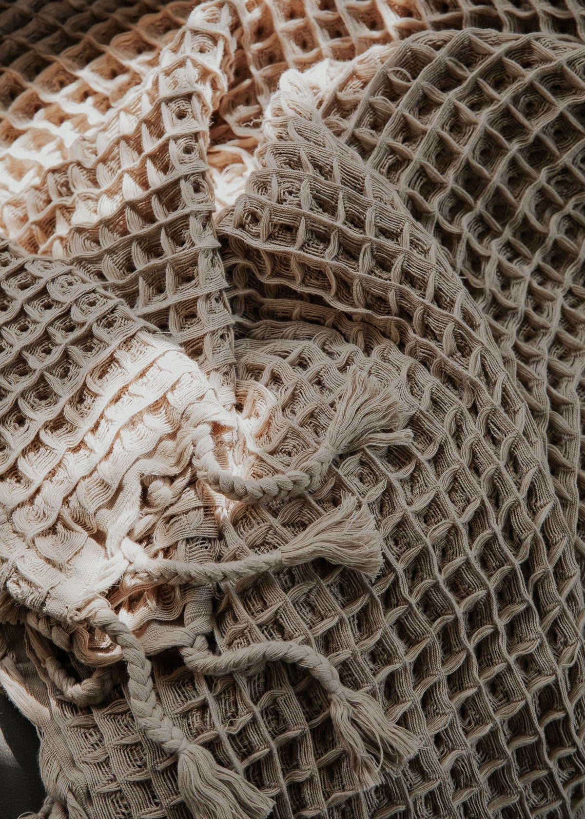 A close-up of a woven throw blanket with a textured grid pattern and tasseled fringe, shown in neutral beige and cream tones.
