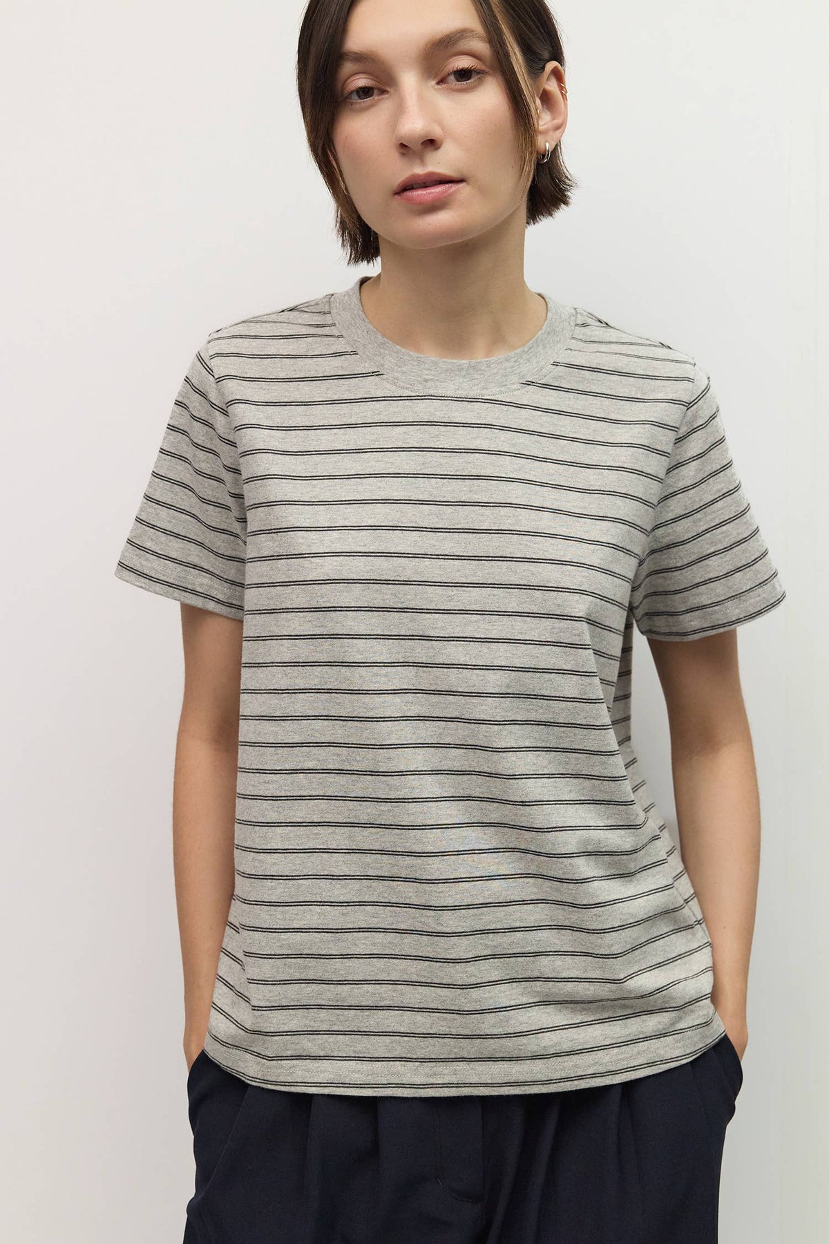 A woman wearing a short-sleeve crewneck t-shirt with thin horizontal gray stripes on a light beige fabric, paired with dark navy pants, against a plain light gray background.