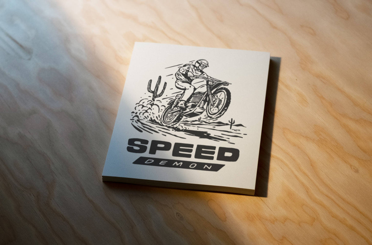 A square white coaster or tile printed with a black line drawing of a motocross rider and the words “SPEED DEMON,” placed on a light wood surface with warm sunlight and shadows.