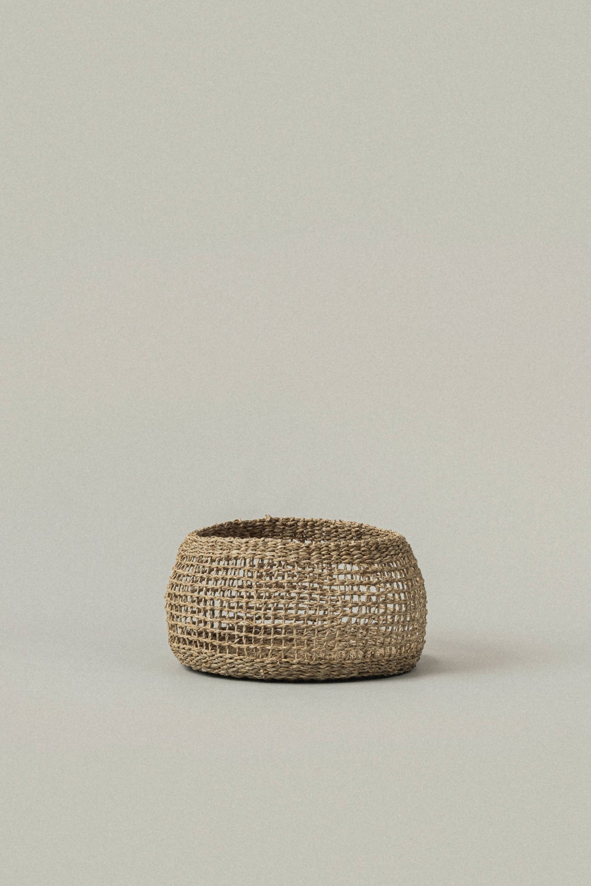 A small round woven basket made from natural jute or seagrass, photographed against a solid, light warm-gray background.