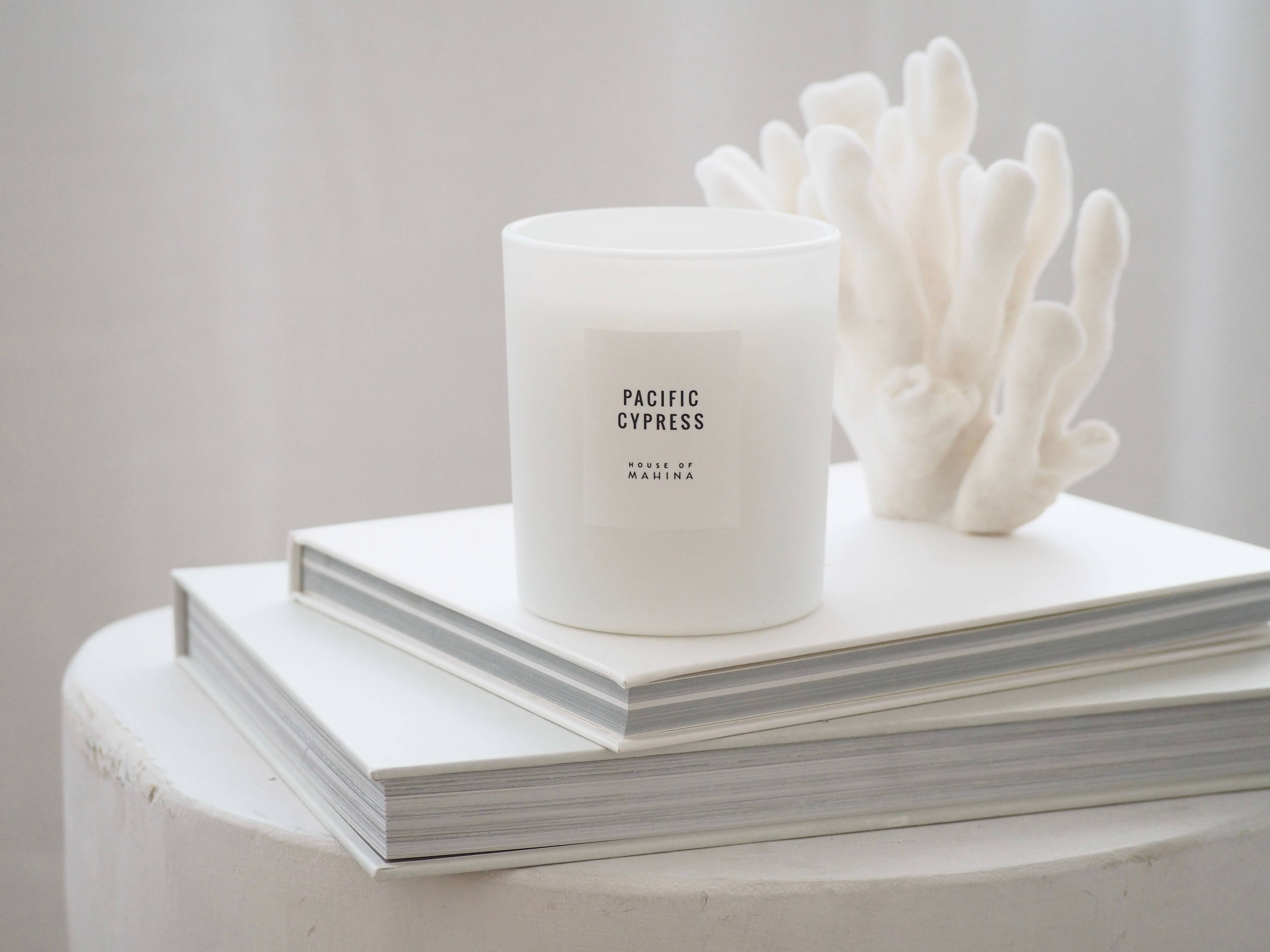 White candle labeled 'Pacific Cypress' on a stack of books with a white coral sculpture.