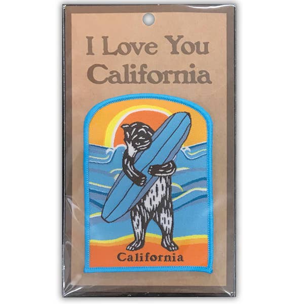 An embroidered patch featuring a bear carrying a blue surfboard on a beach sunset background, mounted on a brown backing card that reads 'I Love You California' and is sealed in clear plastic packaging.