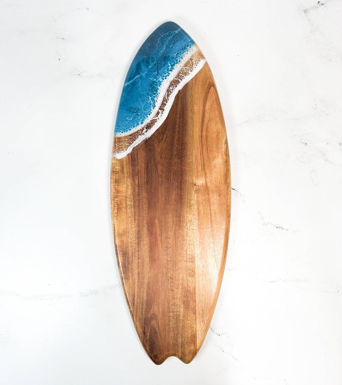 A surfboard-shaped wooden serving board with a blue ocean wave and white foam resin inlay along the top edge, displayed on a white marble surface.
