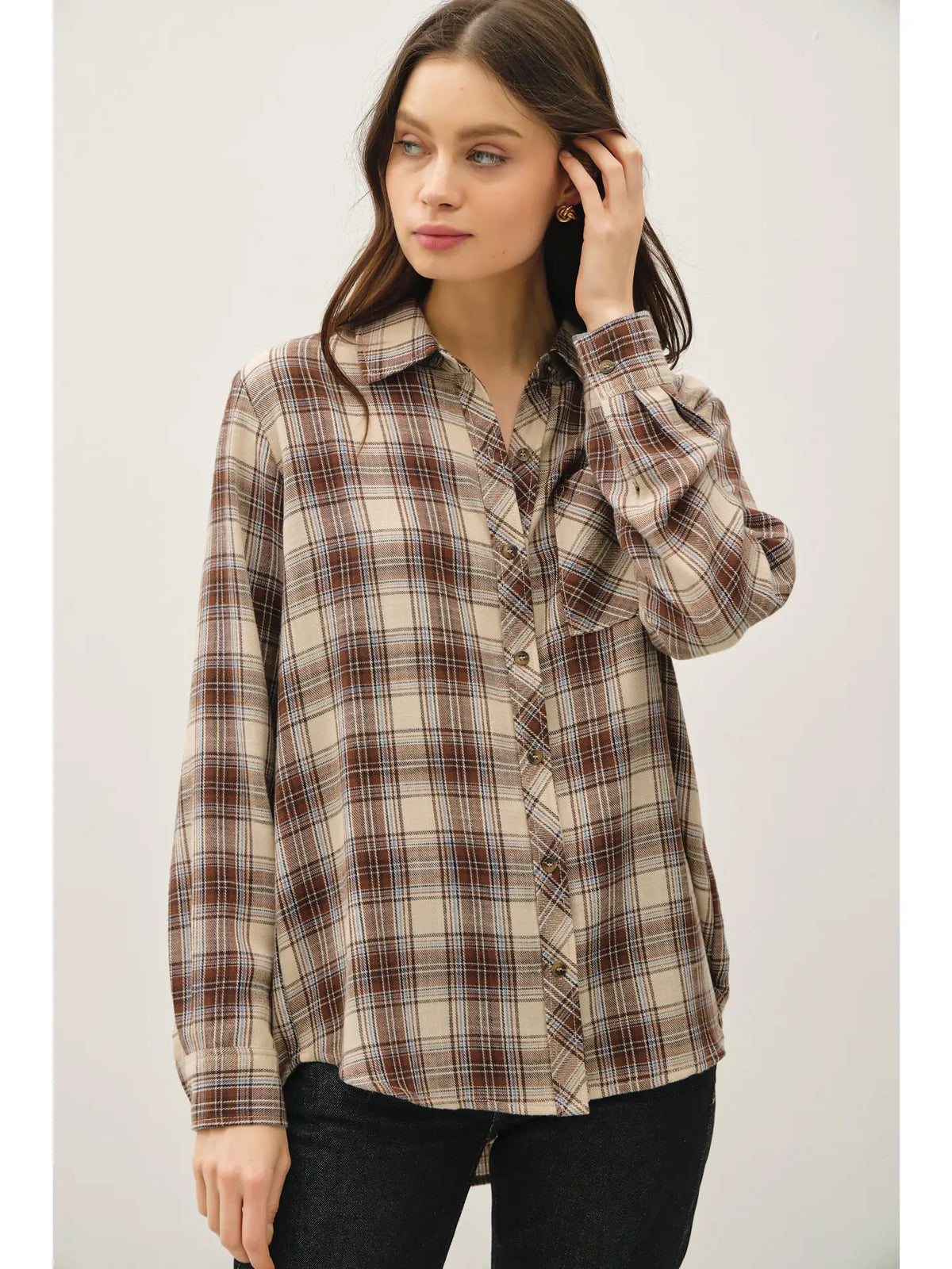 A woman wearing a long-sleeve button-up plaid shirt in beige, brown, and white, paired with black jeans, against a plain light gray background.