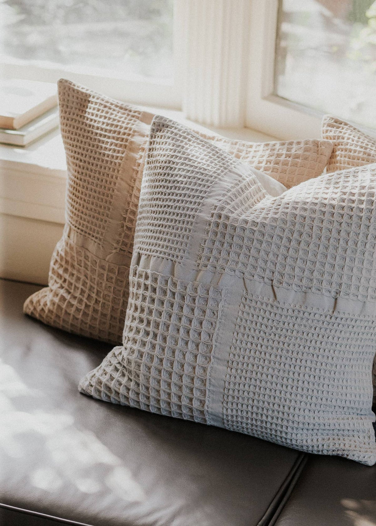 Two textured square throw pillows in warm beige and cool light gray waffle-weave fabric, styled on a dark leather sofa in front of a softly lit window.