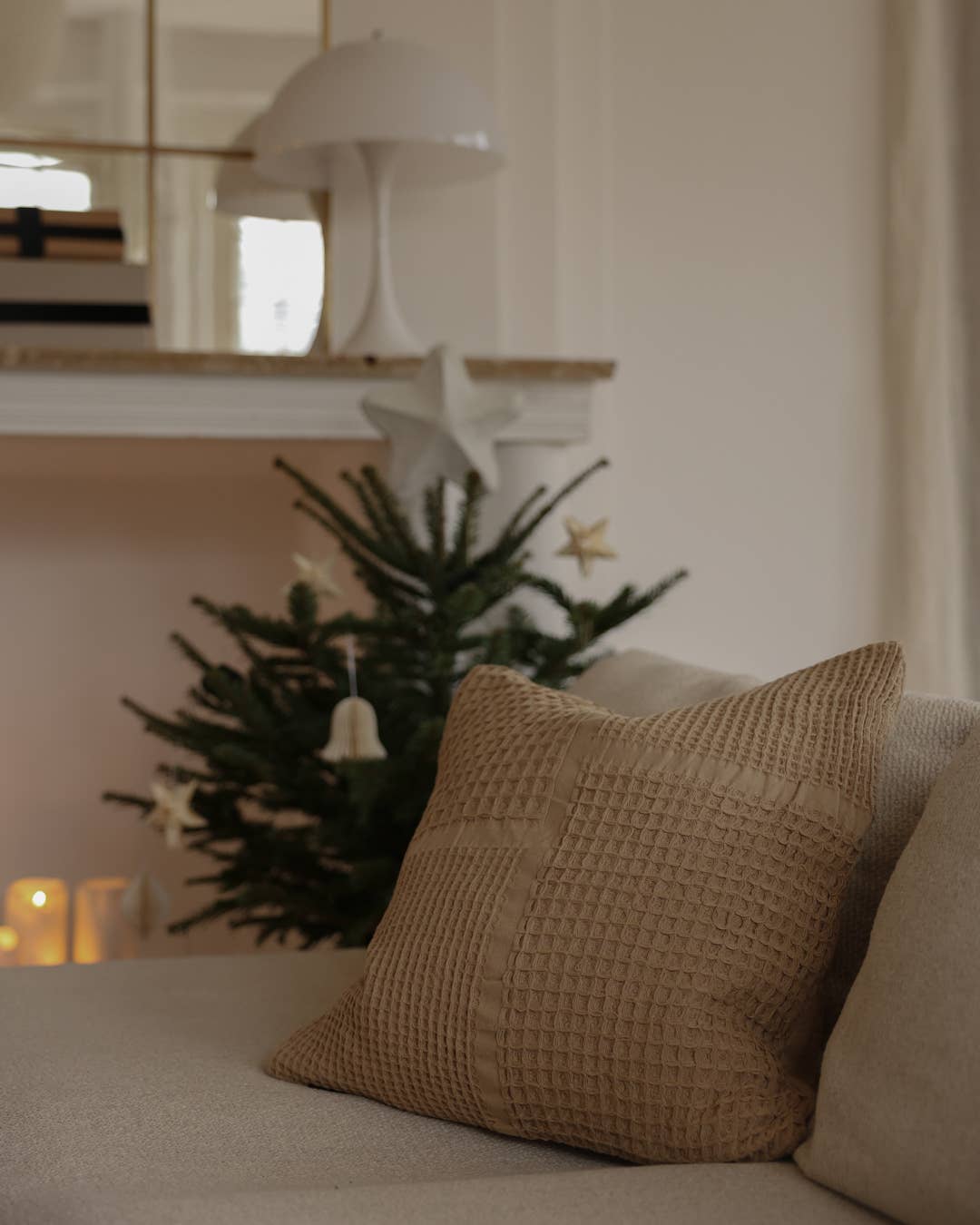 A close-up of a textured beige throw pillow on a light-colored sofa, with a decorated Christmas tree and a white lamp blurred in the background.