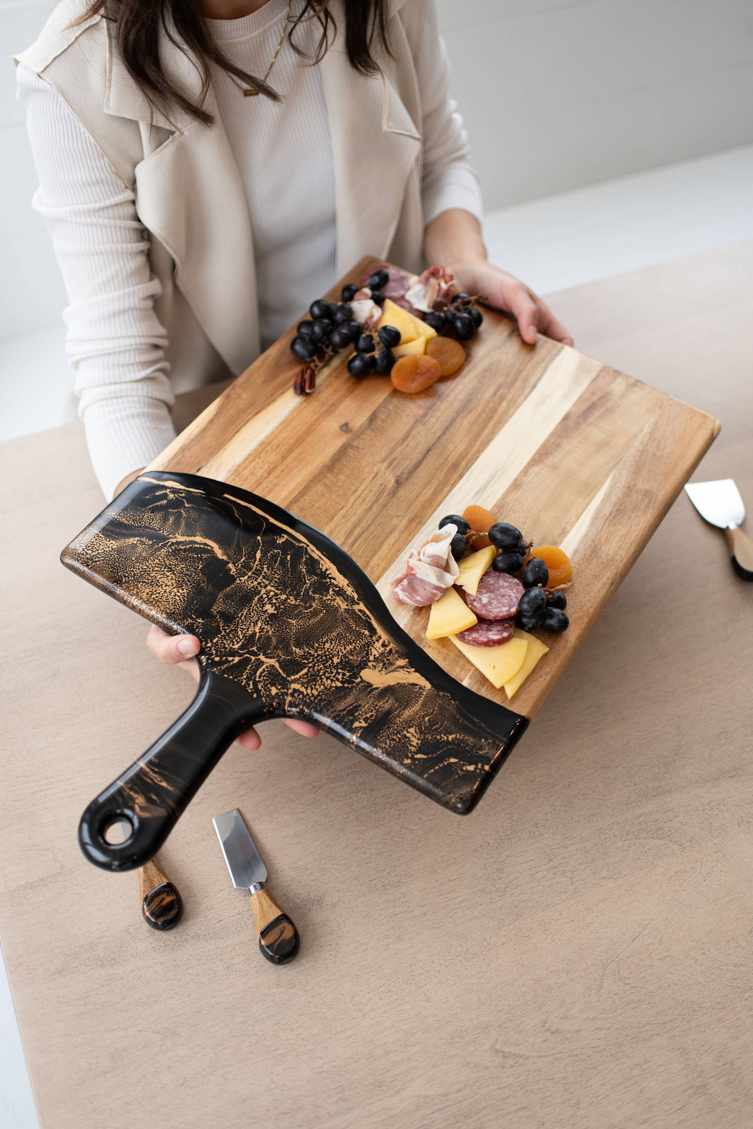 Person holding a wooden cutting board with cheese and fruit, accompanied by a decorative knife.