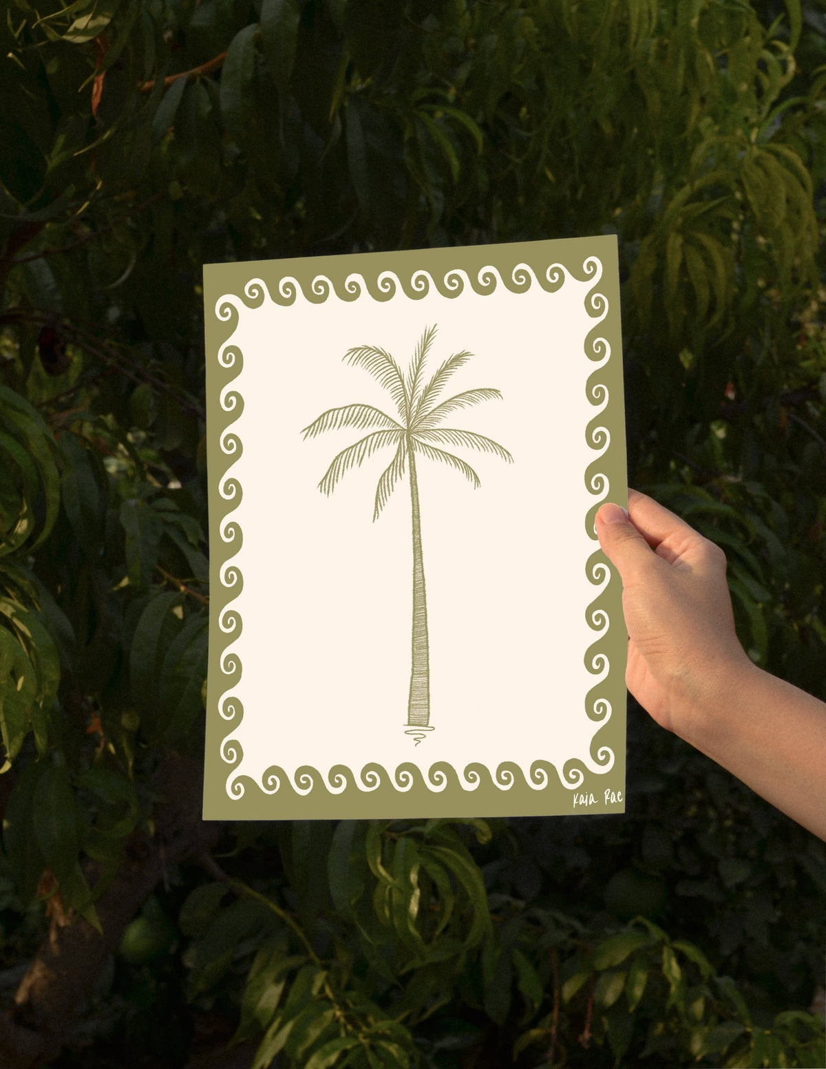 A hand holds a square art print featuring a palm tree illustration with a decorative border, set against a background of green leaves.