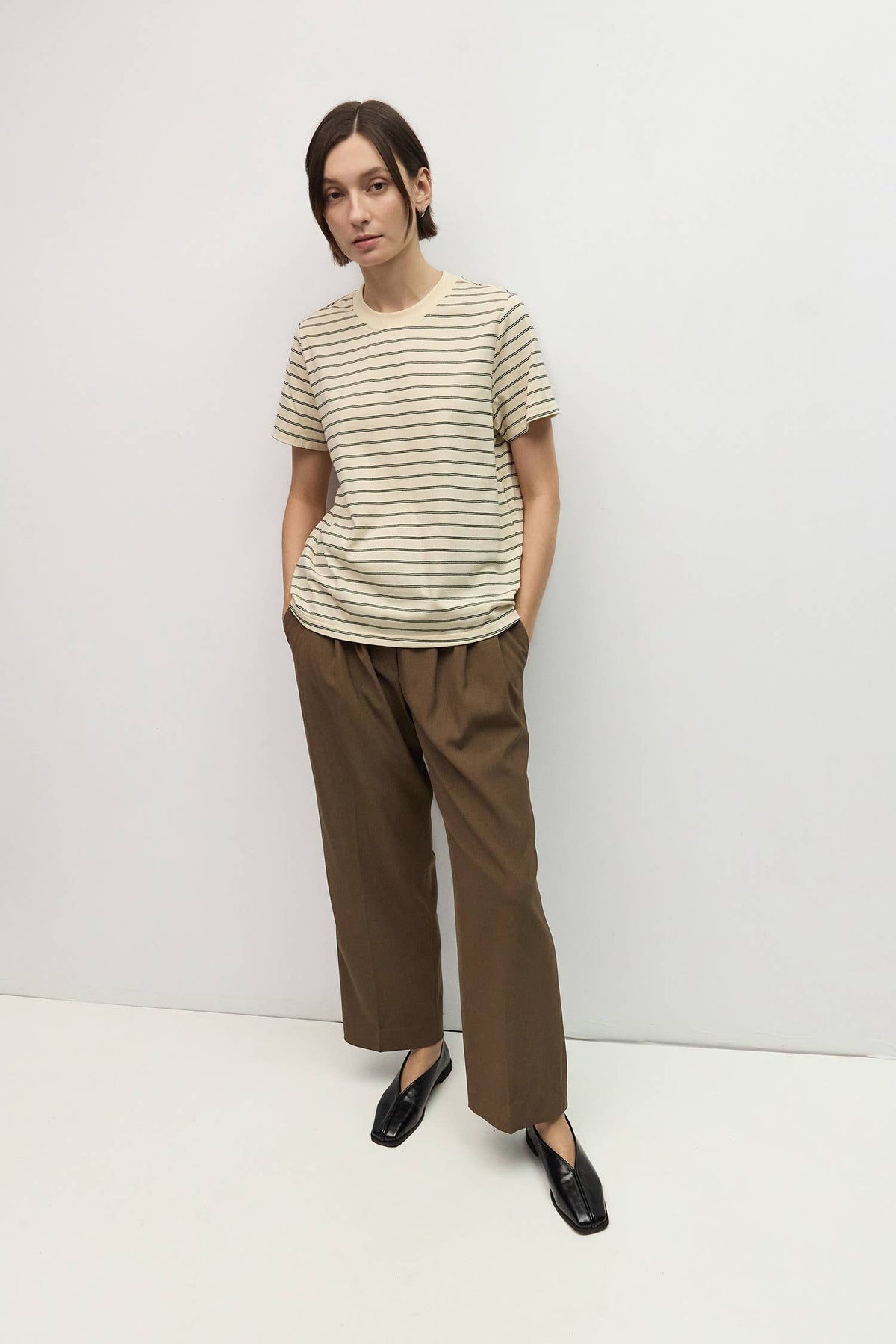 A woman modeling a beige short-sleeve t-shirt with thin dark horizontal stripes and brown wide-leg cropped pants, paired with black loafers, against a plain white studio background.