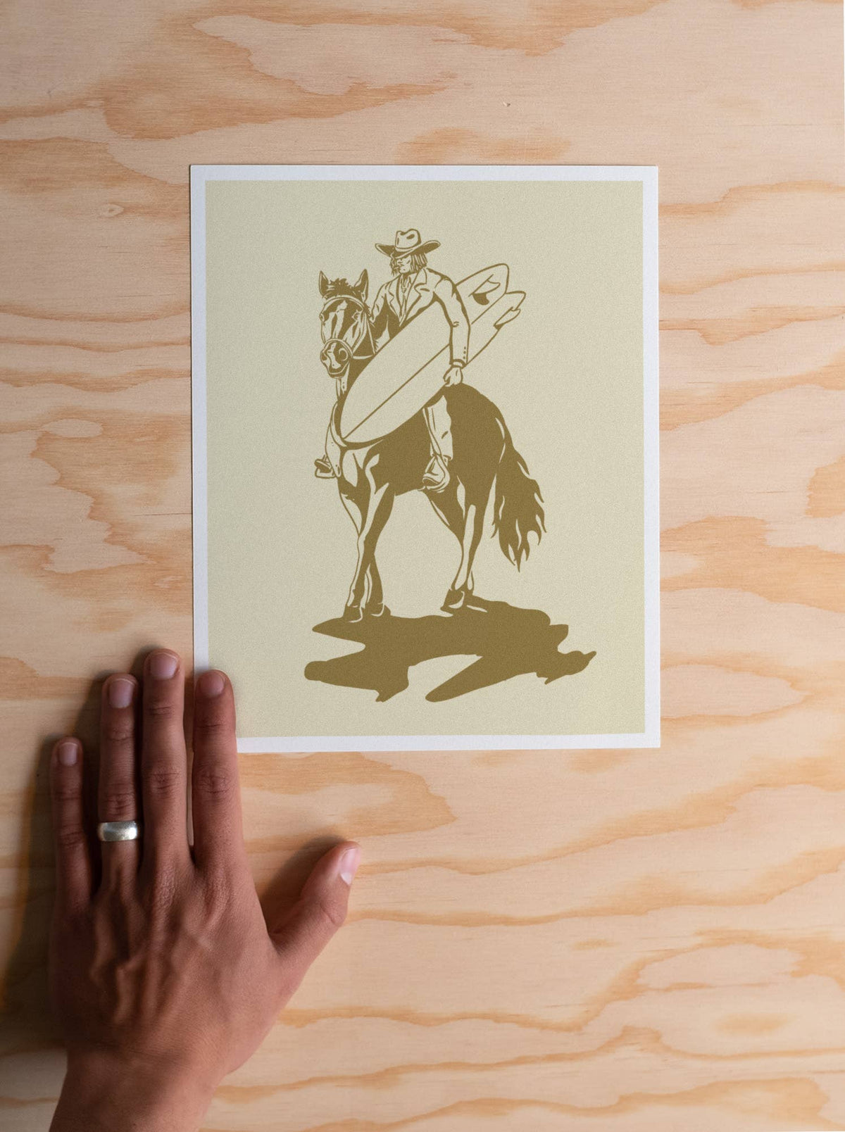 A square art print featuring a minimalist illustration of a cowboy riding a horse while holding a surfboard, displayed on a light wood grain surface with a hand resting underneath the print.