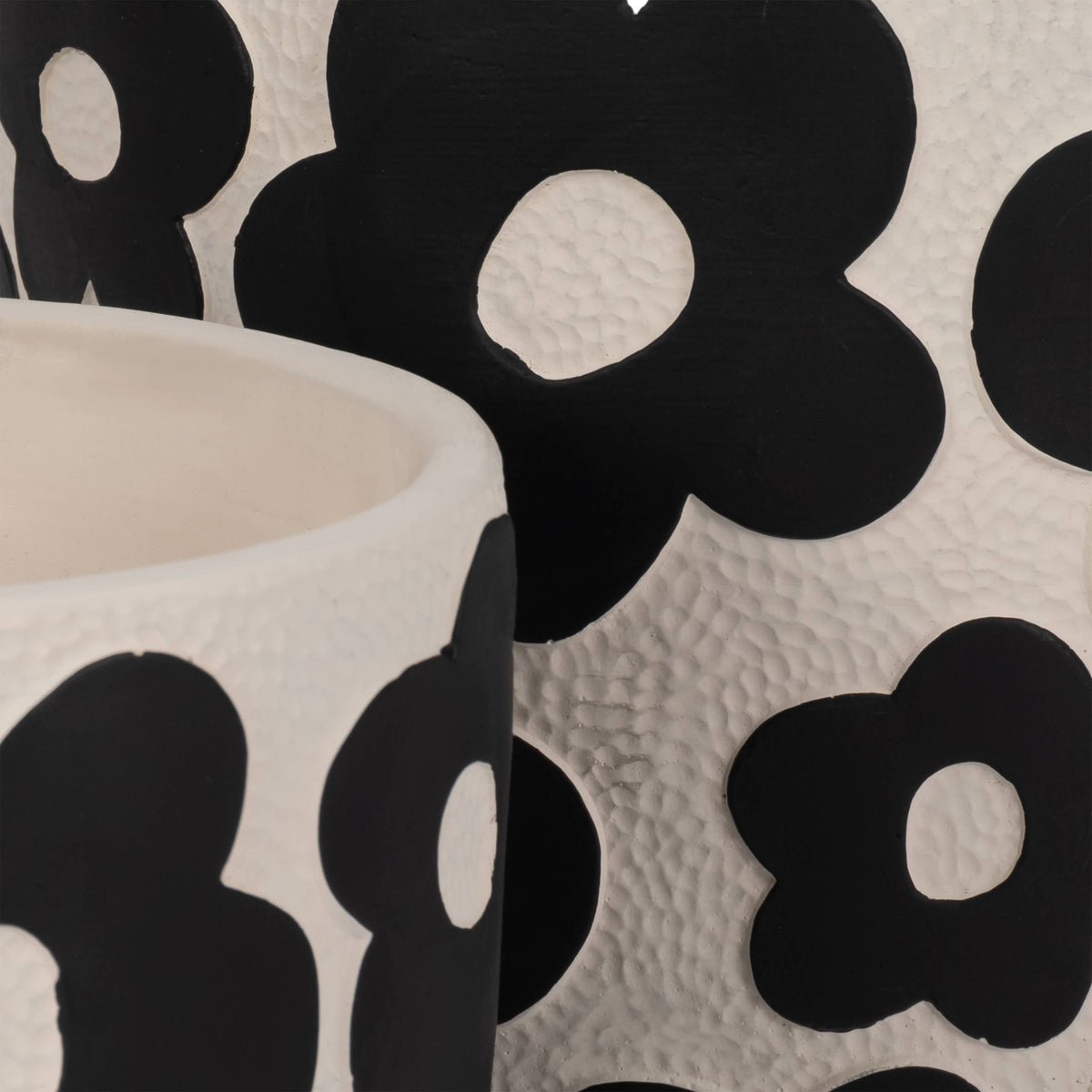 A close-up of a white textured ceramic vessel, likely a candle jar or small container, featuring a bold black floral pattern with flower-shaped cutouts on a matching patterned background.