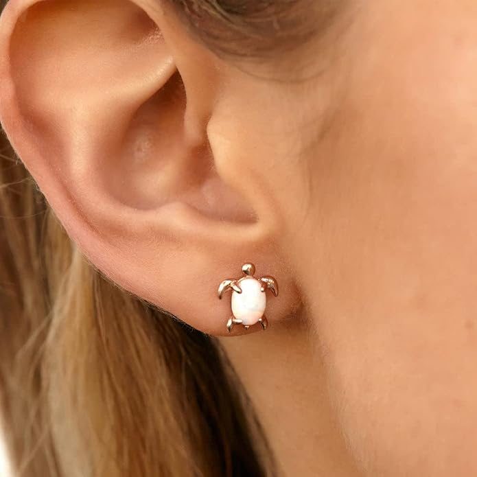 A close-up of a person's ear wearing a small turtle stud earring with a white oval center and rose gold-toned outline.