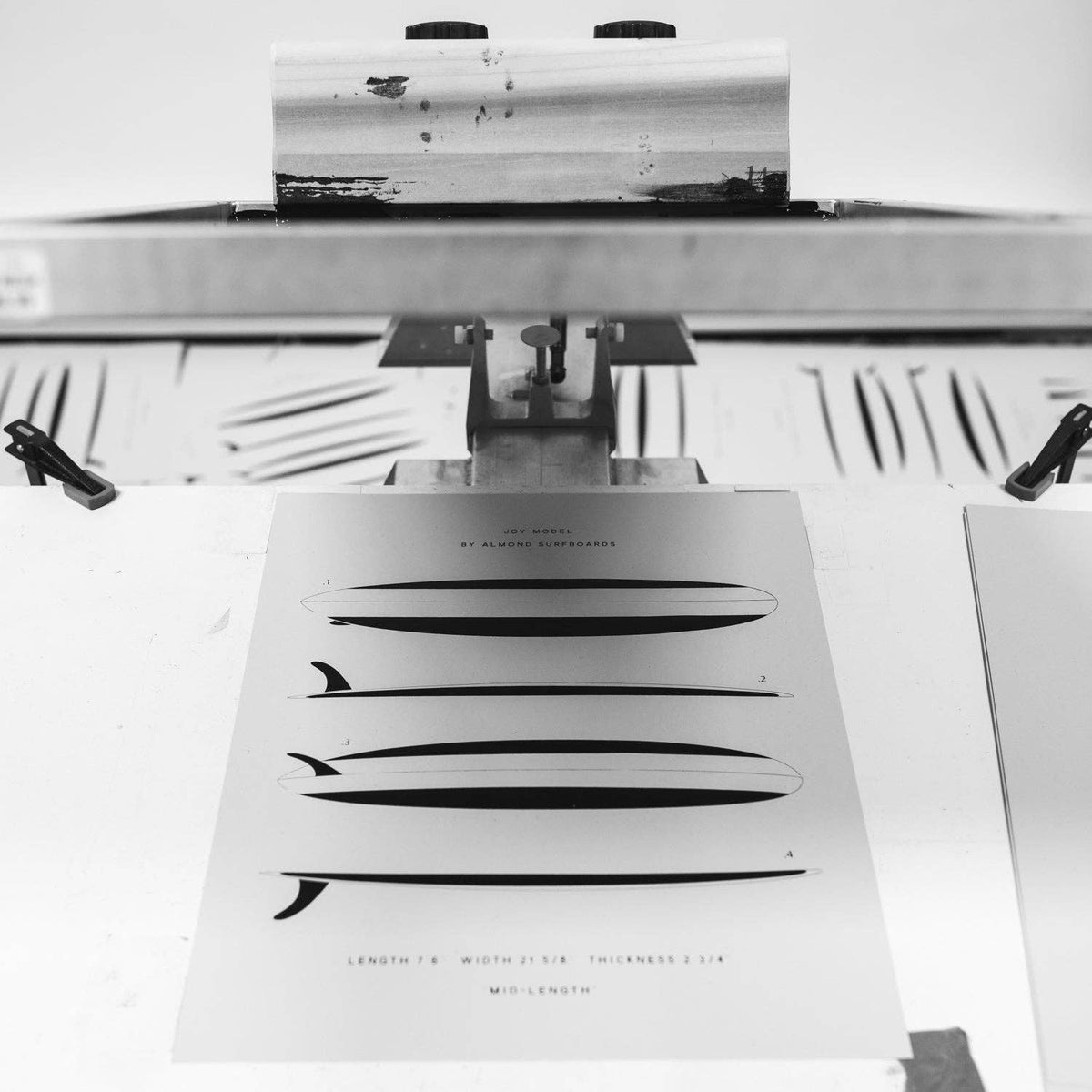A black and white photograph of a surfboard print being pressed onto a sheet by a metal hot stamping press. The print features minimalist graphic designs of various surfboards with text details.