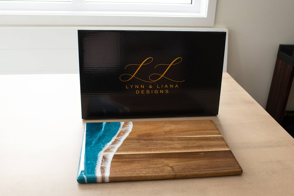 Wooden cutting board with blue resin design on a white surface, branded 'Lynn & Liana Designs'.