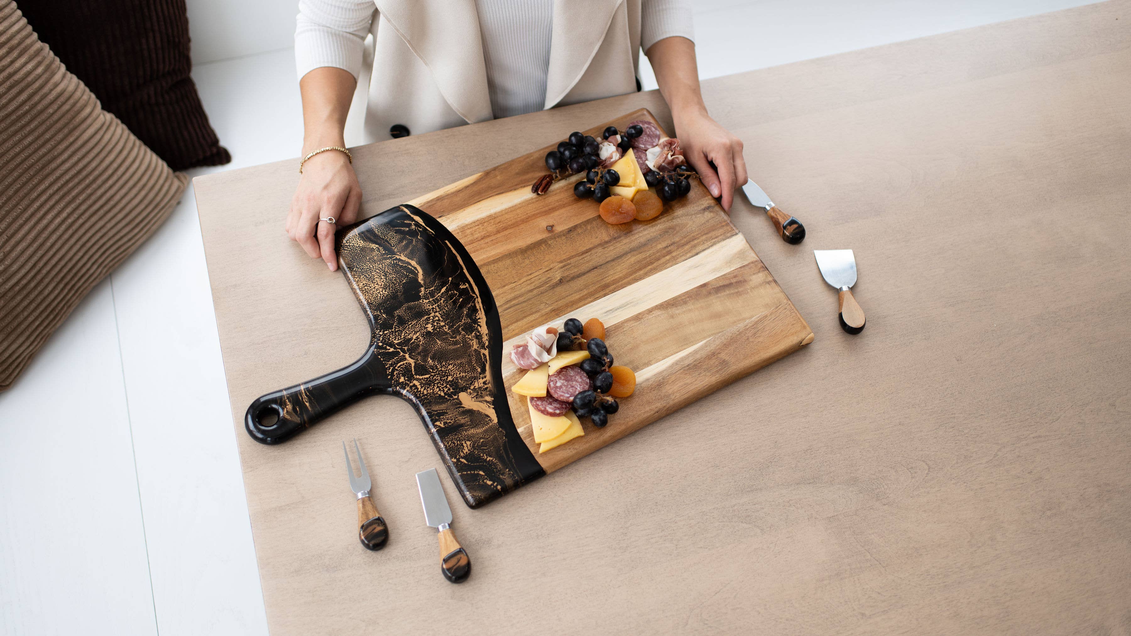 Person arranging food on a wooden cutting board with utensils on a light-colored surface.