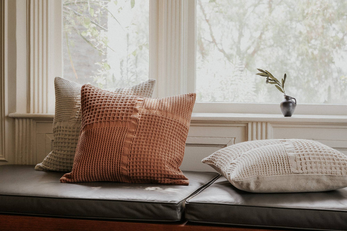 Three textured throw pillows in terracotta, beige, and cream are arranged on a dark leather window seat in a bright room.
