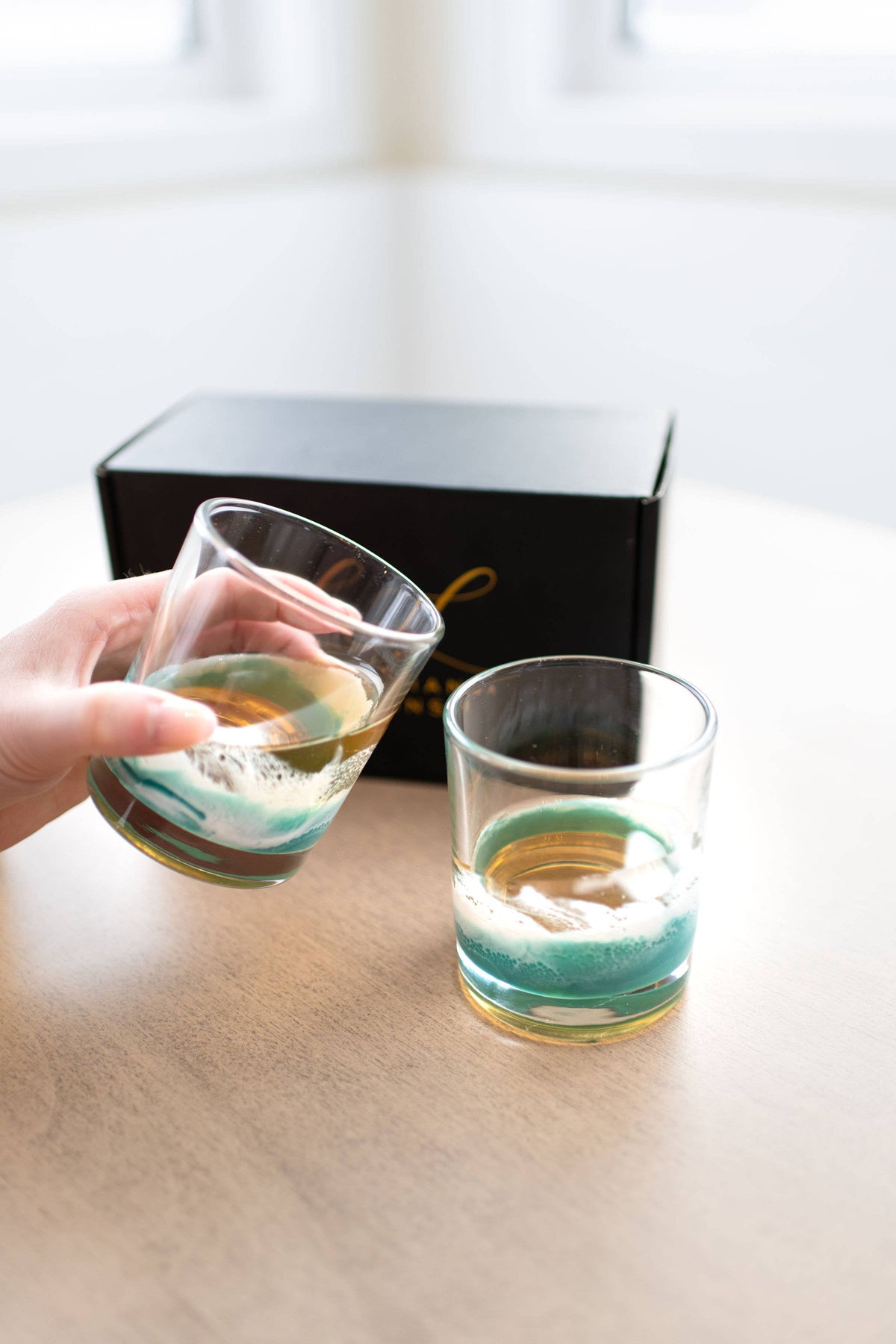 Two glasses with green and gold designs on a wooden surface, one being held by a hand.