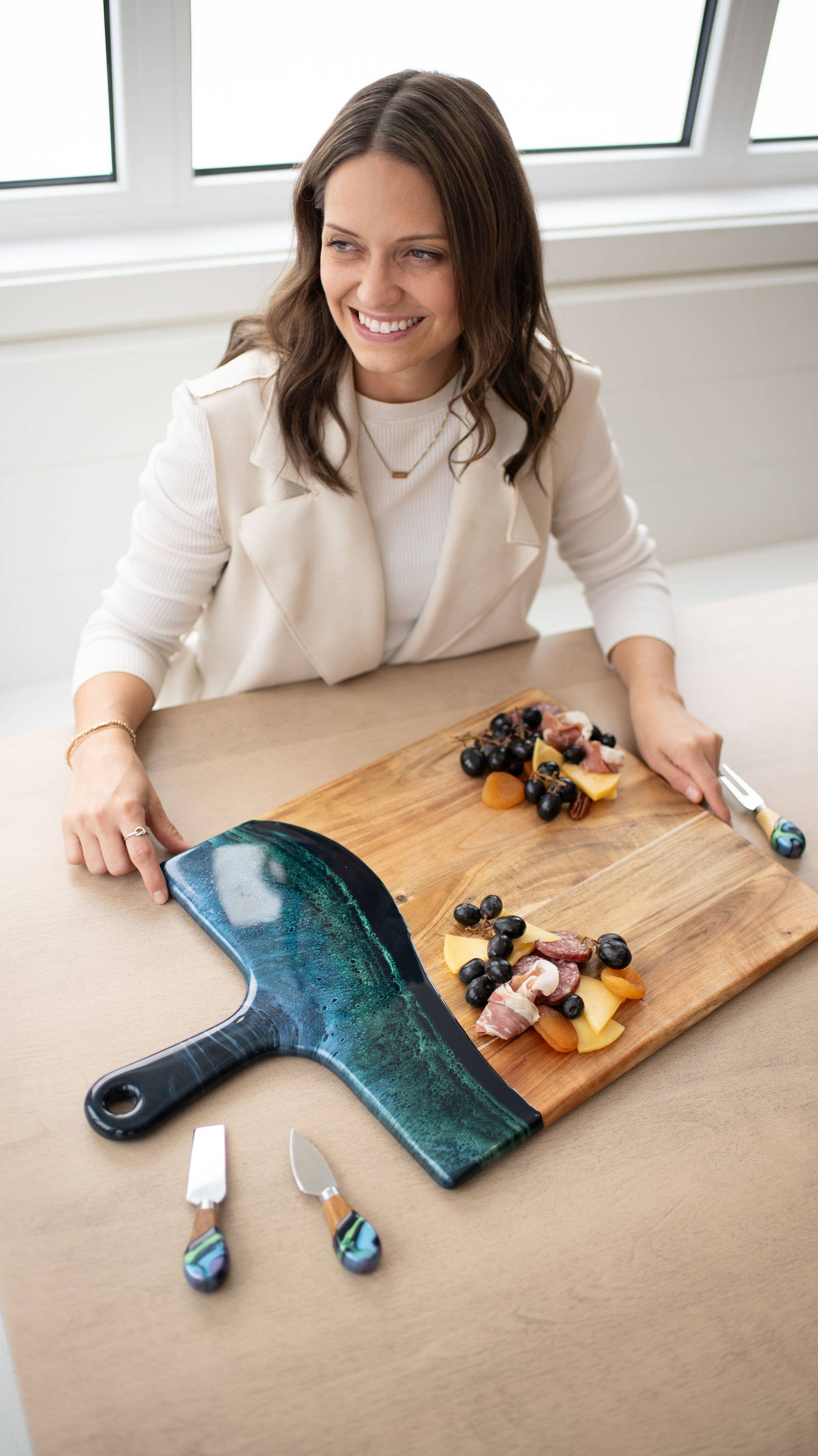 Woman with a wooden cutting board featuring food and a blue cheese knife in a bright room.