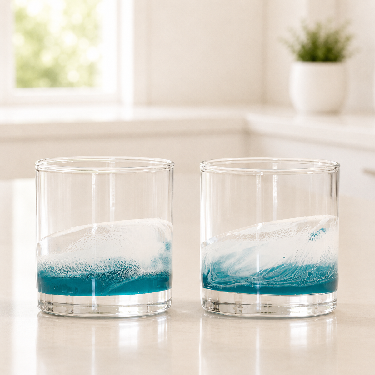 Two clear glass tumblers with a wave design, filled with blue and white liquid, sitting on a light countertop in a bright kitchen setting.