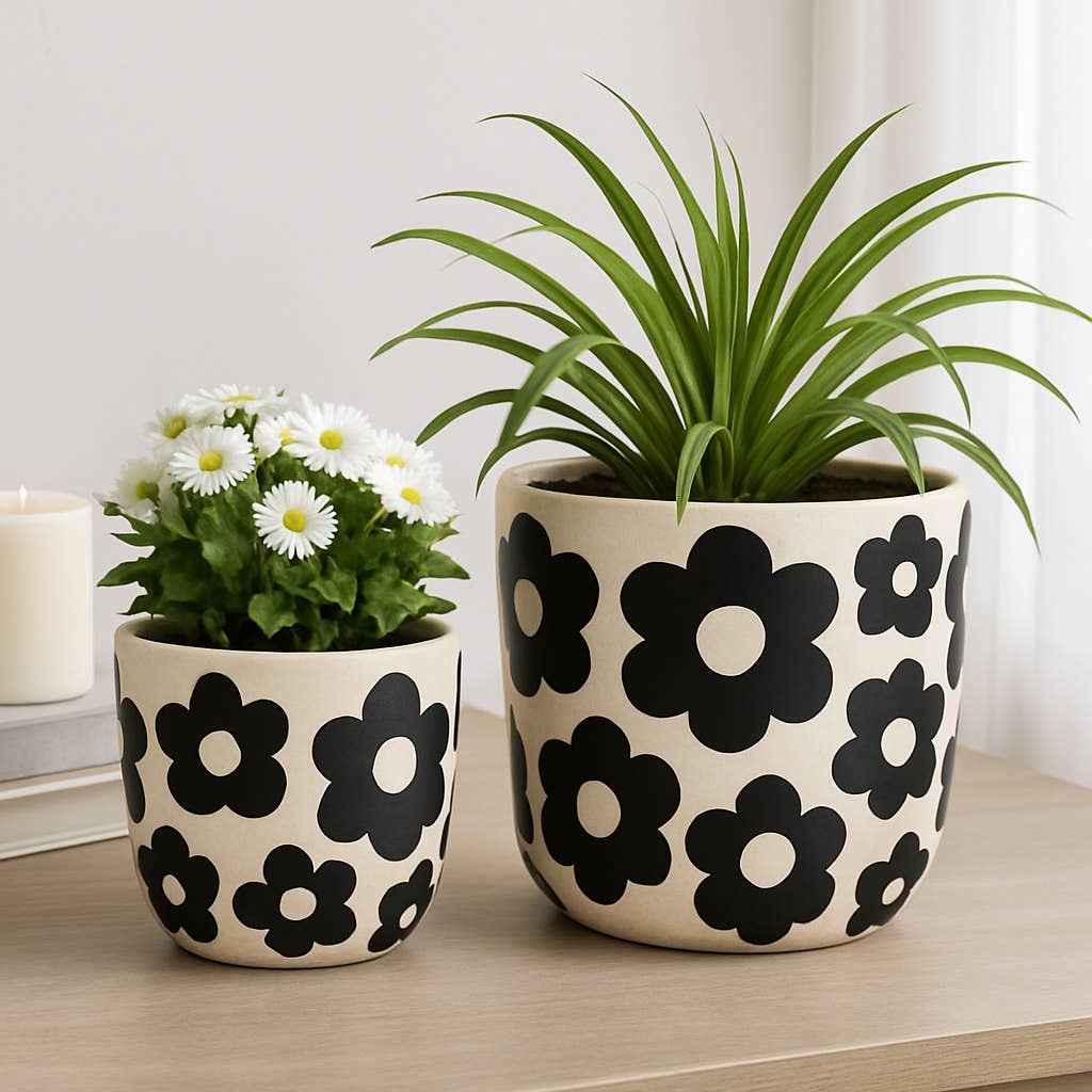 Two ceramic planters with a black floral pattern on a cream background, displayed on a light wood surface. The larger planter holds a green leafy plant, and the smaller planter holds white daisy flowers.